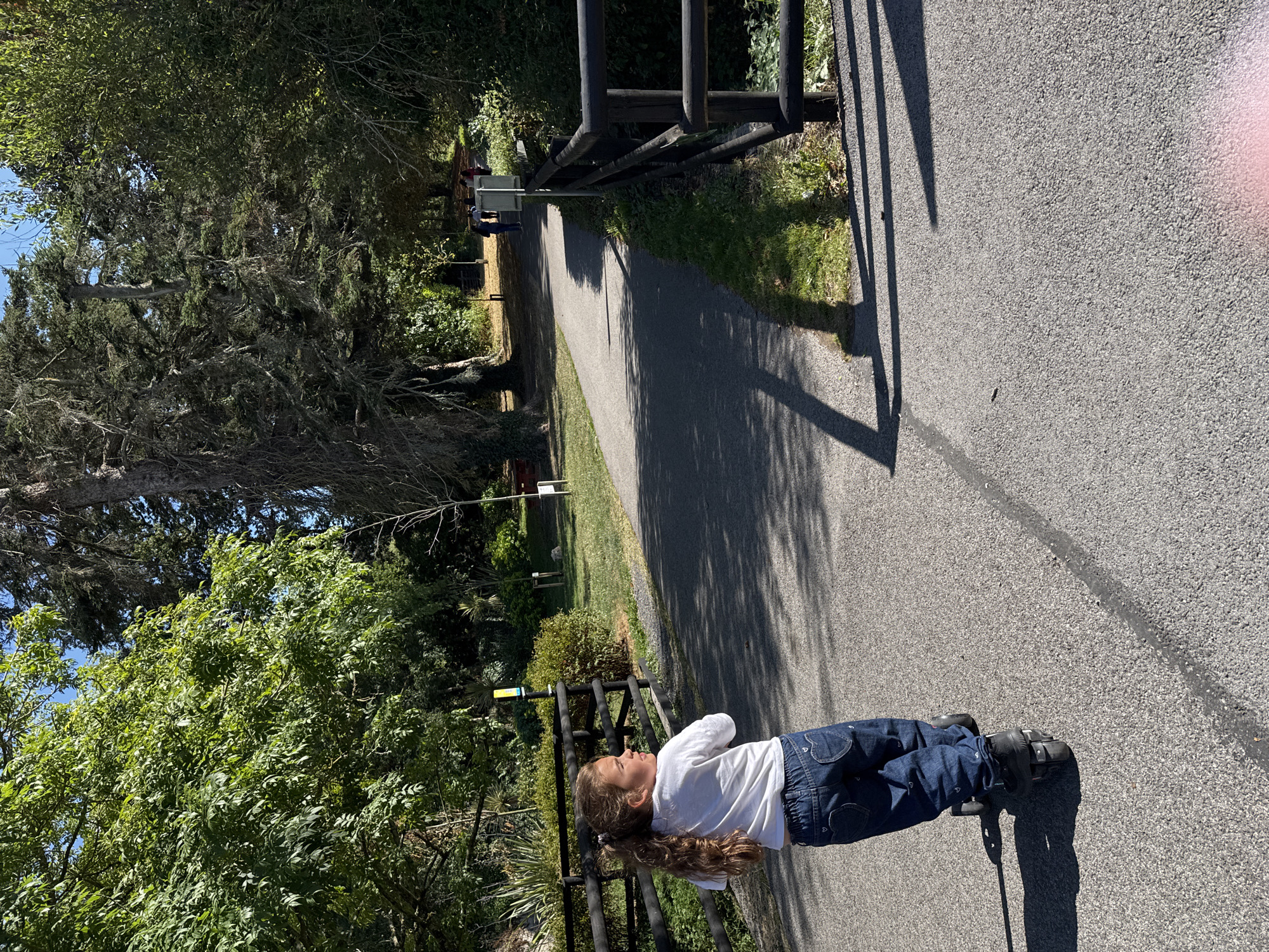 In Cahir Park Gardens, Ireland, a young girl with long, dark hair pulled into a ponytail rides a scooter on a paved path. She wears a white long-sleeved shirt and blue jeans with heart details on the back pockets. The path is bordered by a dark wooden fence on the left and a similar fence on the right, with lush green grass and trees on either side. Sunlight casts shadows on the path. In the background, other people can be seen walking further down the path, and mature trees and foliage create a natural, park-like setting. The sky is visible and appears to be clear and blue.