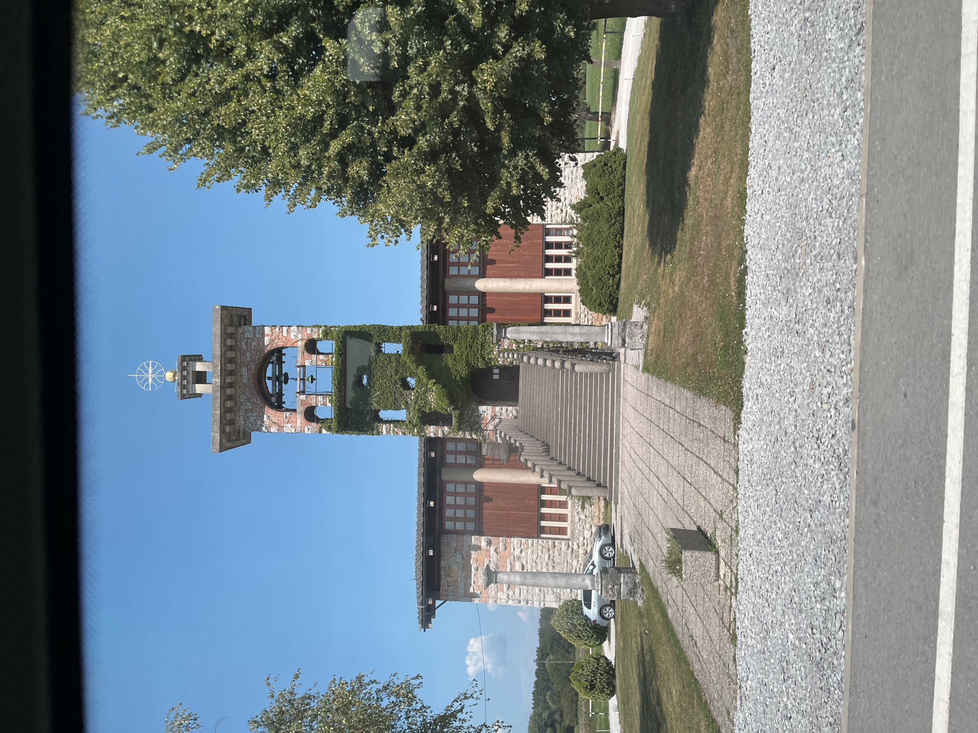 The image shows the Church of the Holy Spirit in Črna vas, Slovenia. It is a bright, sunny day with a clear blue sky. The church has a distinct bell tower covered in ivy, with a golden dome and a cross at the very top. In front of the bell tower is a large set of stairs leading up to the entrance of the church. The church building itself is made of stone and wood, with large windows. To the left of the church, a silver car is parked on a paved pathway. The area around the church is grassy, with some bushes and trees. The foreground shows a gravel path and a road with a white line.