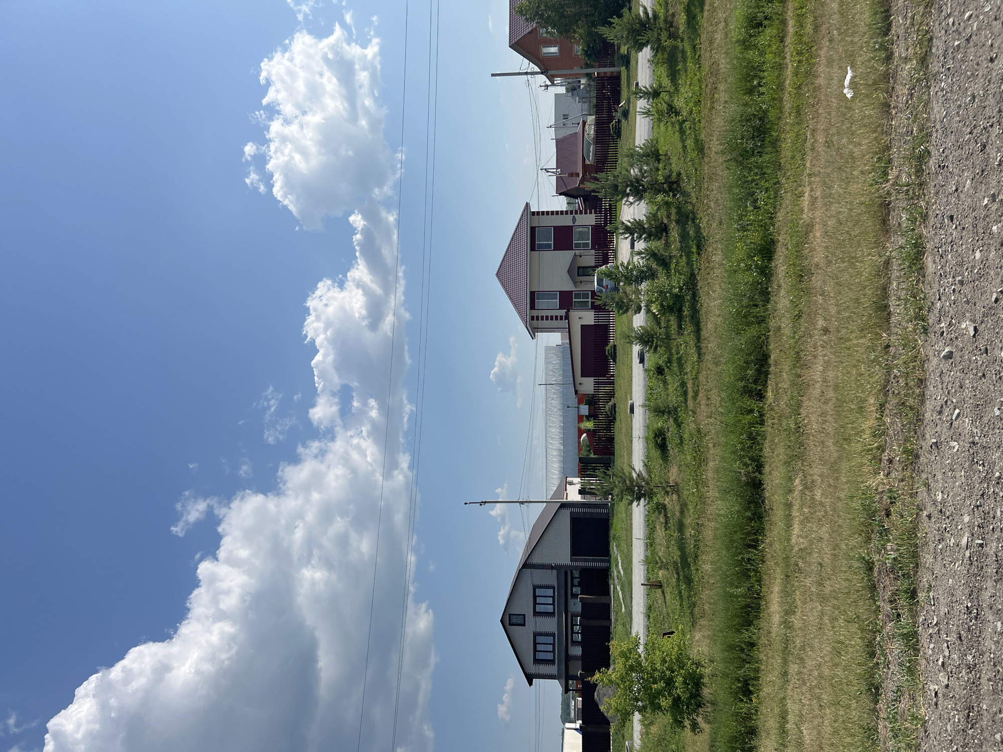 The image depicts a suburban street scene in Prokudskoye, Russia, on a clear, sunny day. The foreground is dominated by a gravel road and a strip of dry, sparse grass. Beyond this, a line of young evergreen trees runs parallel to a paved road.
Along the paved road, several residential houses are visible, separated by wooden fences. The houses are of various designs and colors, with pitched roofs. One house on the left is light-colored with dark trim and a dark garage. To its right, a larger, two-story house features a light-colored facade with maroon accents and a maroon roof. Further to the right, another house has a brick facade and a red roof. In the background, larger industrial-looking structures and more houses are present.
The sky is a bright blue with scattered white cumulus clouds. Power lines stretch across the upper portion of the image. The overall impression is one of a developing residential area on the outskirts of a town, characterized by new construction and open spaces.