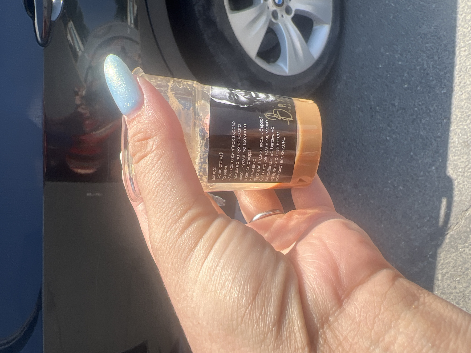 A person's hand with light blue, glittery fingernails holds a clear plastic cup of coffee. The cup has text printed on it in Bulgarian, which appears to be a joke about a doctor and patient. A portion of a dark-colored car, including its wheel and door handle, is visible in the background. The ground appears to be asphalt. The image was taken outdoors in Burgas, Bulgaria.