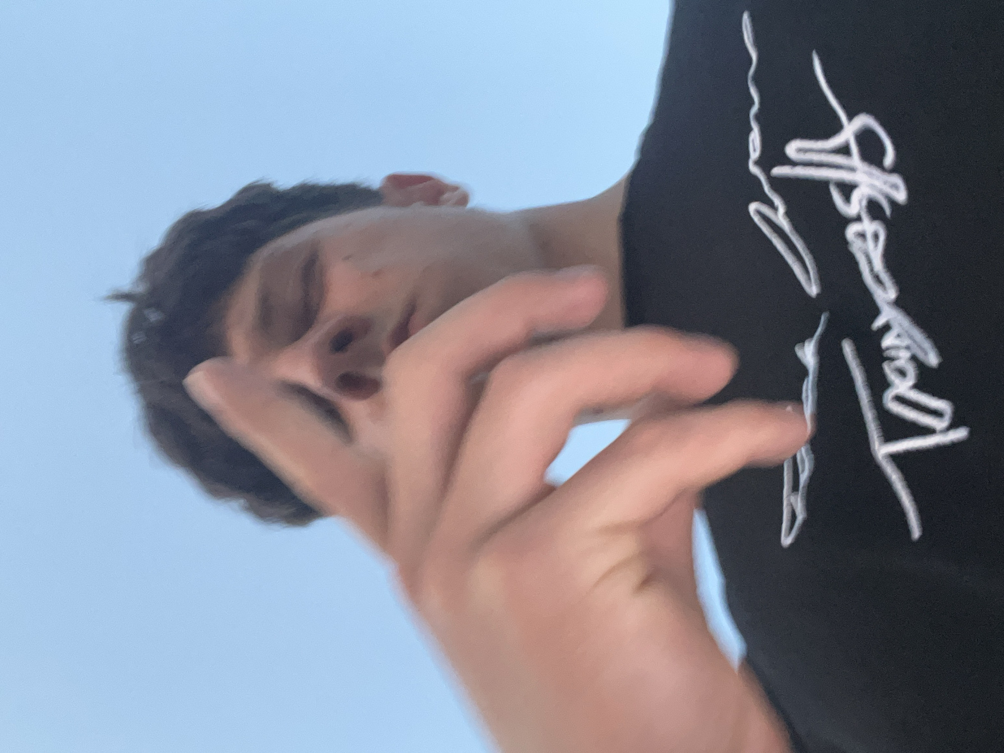 A low-angle, upward-facing photographic image depicts a single male individual under a clear, light blue sky, presumably outdoors in the city of Zlatograd, Bulgaria. The subject's head is positioned towards the upper center of the frame, with dark, short hair visible. His eyes appear closed or directed downwards, and his facial features are partially obscured by his right hand (viewer's left), which is raised into the foreground, significantly out of focus due to its proximity to the camera. The index finger of this hand is extended, pointing generally towards the camera. The subject is wearing a dark, likely black, short-sleeved t-shirt. White, stylized text is embroidered or printed on the front of the shirt, appearing to read "Train Costs" with additional calligraphic elements. The overall lighting is bright and even, consistent with daylight conditions.