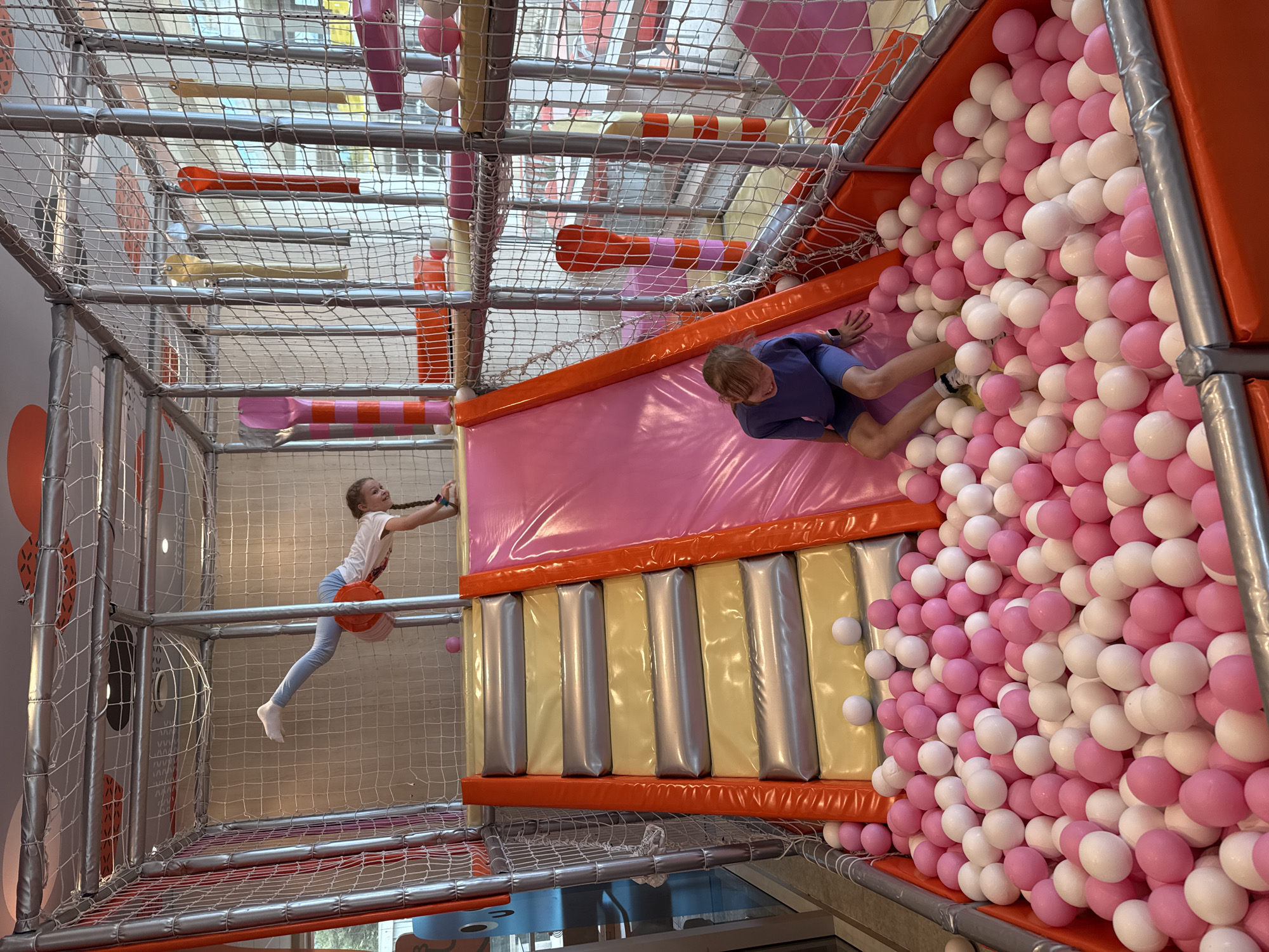 The photograph depicts a children's indoor playground, likely located in a mall or entertainment center. The scene is viewed from an elevated, slightly tilted perspective, showcasing the various play structures and activities.
A young girl with pigtails, wearing a white t-shirt and light-colored pants, is suspended mid-air while holding onto the side of a climbing structure or net. She appears to be in motion, possibly swinging or climbing.
A young boy, clad in a blue outfit, is positioned inside a pink slide leading into a ball pit. He is shown near the transition point between the slide and the ball pit, where numerous pink and white balls are densely packed.
The playground features a metal framework overlaid with netting to ensure safety. Various padded structures, including a pink slide, orange borders, and padded steps, create the play area. Overhead elements include pipes and decorative features.
The location is not definitively ascertainable from the image alone; however, according to the prompt's request, the location is Budva, Montenegro.