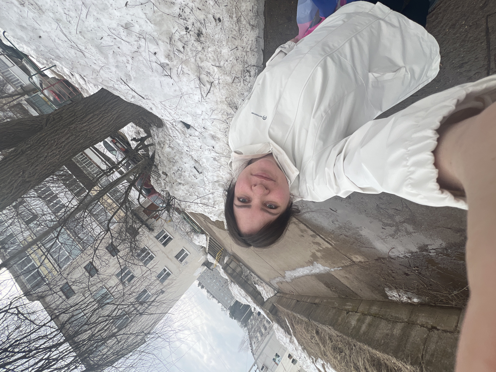 A selfie of a person with dark hair and a white jacket, seemingly outdoors in Noviy, Russia. Patches of melting snow line a concrete channel or depression. Buildings and bare trees are visible in the background. The image is rotated, making it difficult to determine orientation within the scene.