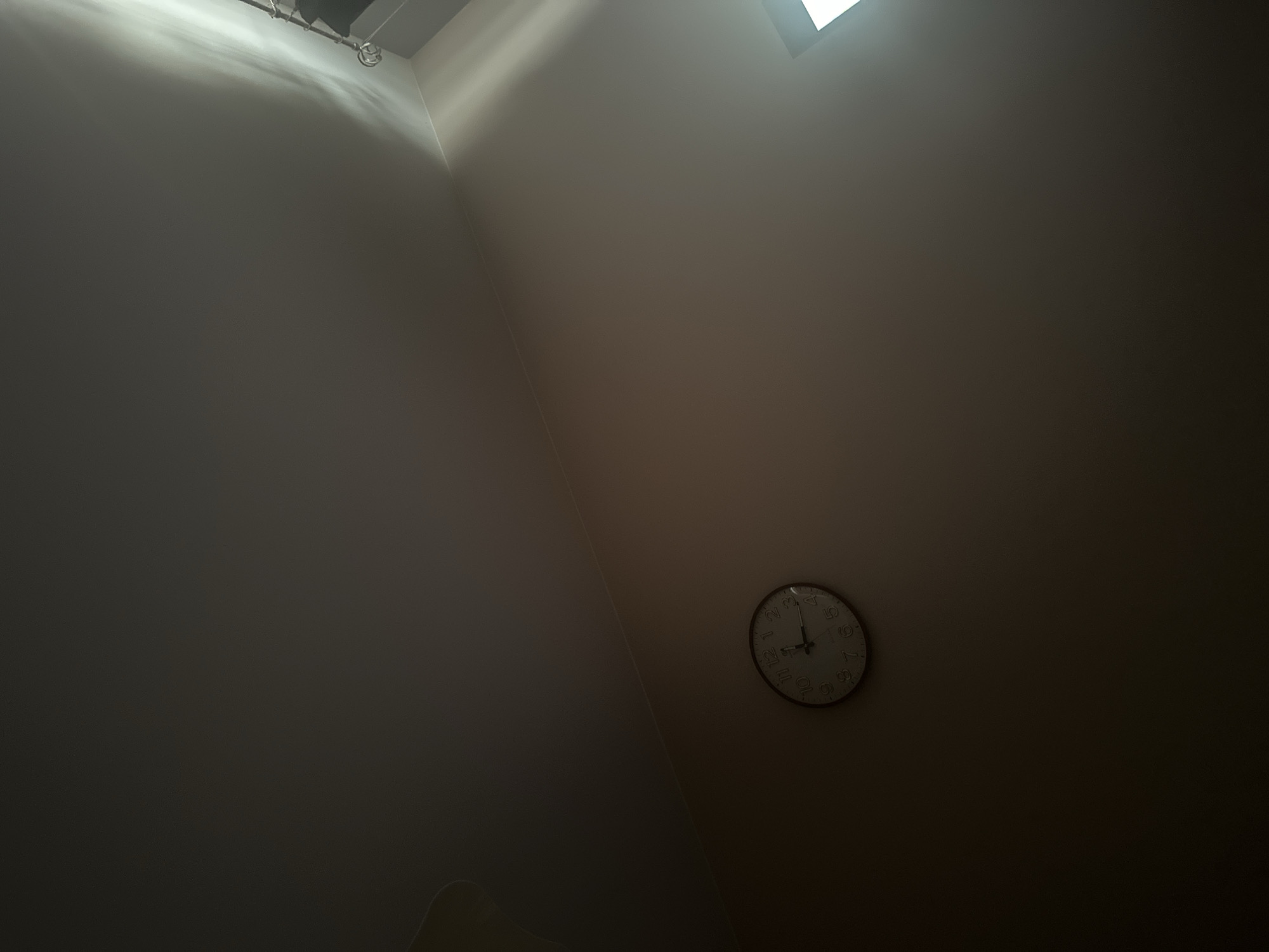 The image shows a close-up view of a round analog clock mounted on a plain, light-colored wall. The clock has a dark bezel and a white face with black numbers and hands. The time displayed on the clock is approximately 10:42. The lighting in the image is dim, with a bright shaft of light entering from the upper right corner, casting shadows and illuminating the upper portion of the wall and ceiling. The scene is indoors, likely a room, with no other objects or people clearly visible. The location is specified as Tuszynek Majoracki, Poland.