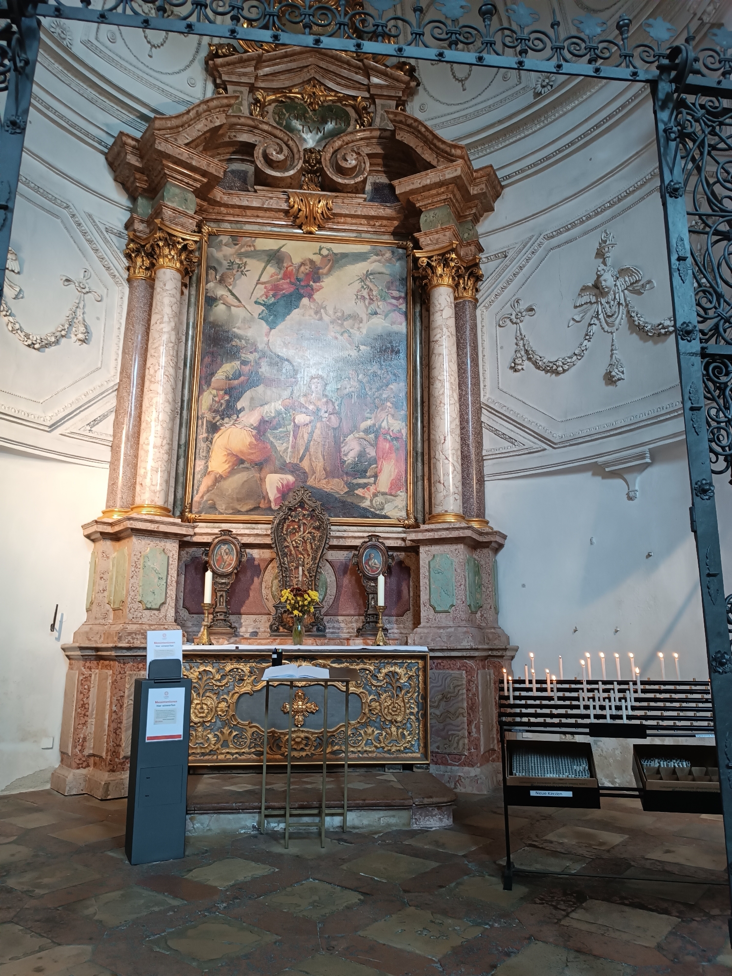 The image depicts the interior of a church in Munich, Germany, likely Saint Michael's Church (Michaelskirche) given the prominent altar with a large painting. The altar is elaborately decorated with marble columns, gilded details, and a central religious painting showing figures in a heavenly or biblical scene with cherubs. In front of the altar, there is a table with candles, a vase of flowers, and decorative elements. To the left of the altar, a dark grey information stand is visible, with a sign on it. To the right, a row of votive candles are lit and arranged in racks, with a box labeled "Neue Kerzen" (New Candles) beneath them. The church architecture features ornate white stucco decorations on the walls and ceiling. The floor is tiled with large, square stones in shades of red and brown. The overall scene is one of solemnity and religious devotion.