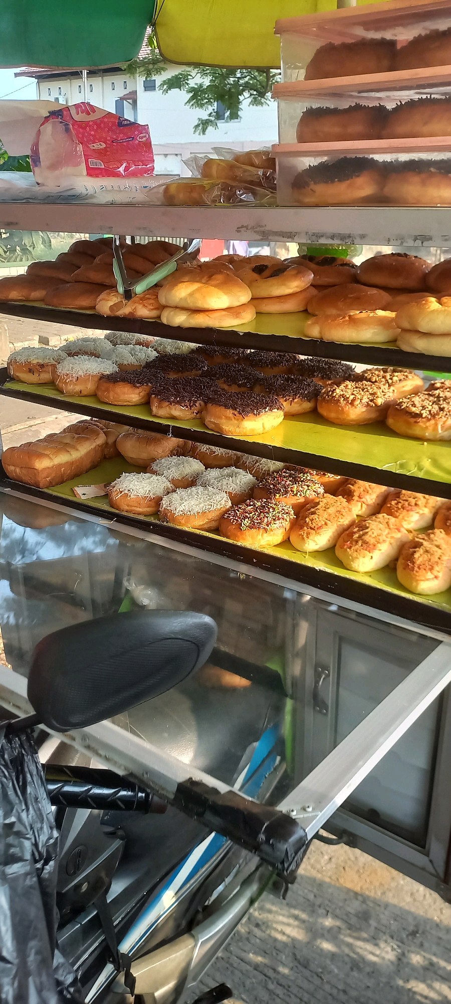 In Melayu, Indonesia, a mobile bakery stall displays an assortment of freshly baked goods. The stall features three shelves laden with bread and donuts, some topped with chocolate sprinkles, shredded cheese, or savory fillings. A pair of green tongs rests on the top shelf, suggesting recent customer service. The stall appears to be set up outdoors, with a hint of greenery and buildings in the background, illuminated by natural light. In the foreground, a portion of a motorcycle, including its seat and handlebars, is visible, indicating the stall may be located on a street or sidewalk. A black plastic bag hangs from the motorcycle's handlebars, and the motorcycle's body shows blue and white decals. The overall scene suggests a vibrant street food vendor operating in a typical Indonesian urban setting.