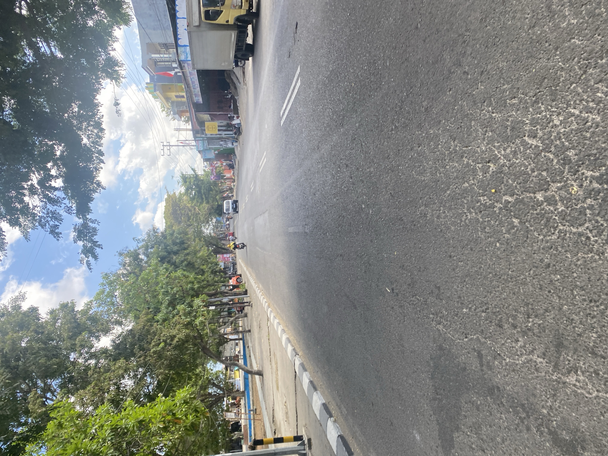 The image depicts a street scene in Bentengnge, Indonesia, on a partly cloudy day. The perspective is from the street level, looking down a paved road lined with trees on the left and buildings on the right.
On the left side of the road, a sidewalk runs alongside a grassy area with scattered trees and some blue-colored public seating. A black and yellow striped pole is visible on the sidewalk. Further down the street on the left, there are more trees and some small structures.
The road itself is asphalt with white double lines marking the lanes. Several vehicles are present. A motorcycle with a rider wearing a yellow helmet is in the middle of the road. In the distance, a white van is approaching. On the right side of the road, there is a row of buildings, some appearing to be shops or businesses with open fronts. A large, light-colored truck is parked in front of one of these buildings. People can be seen sitting or standing in front of the buildings.
The buildings on the right have various colors and architectural styles, with some displaying flags, including the Indonesian flag. Overhead, power lines and trees with green foliage create a canopy over parts of the scene. The sky is blue with scattered white clouds, and sunlight is filtering through the trees. The overall environment suggests a moderately busy urban or semi-urban street.