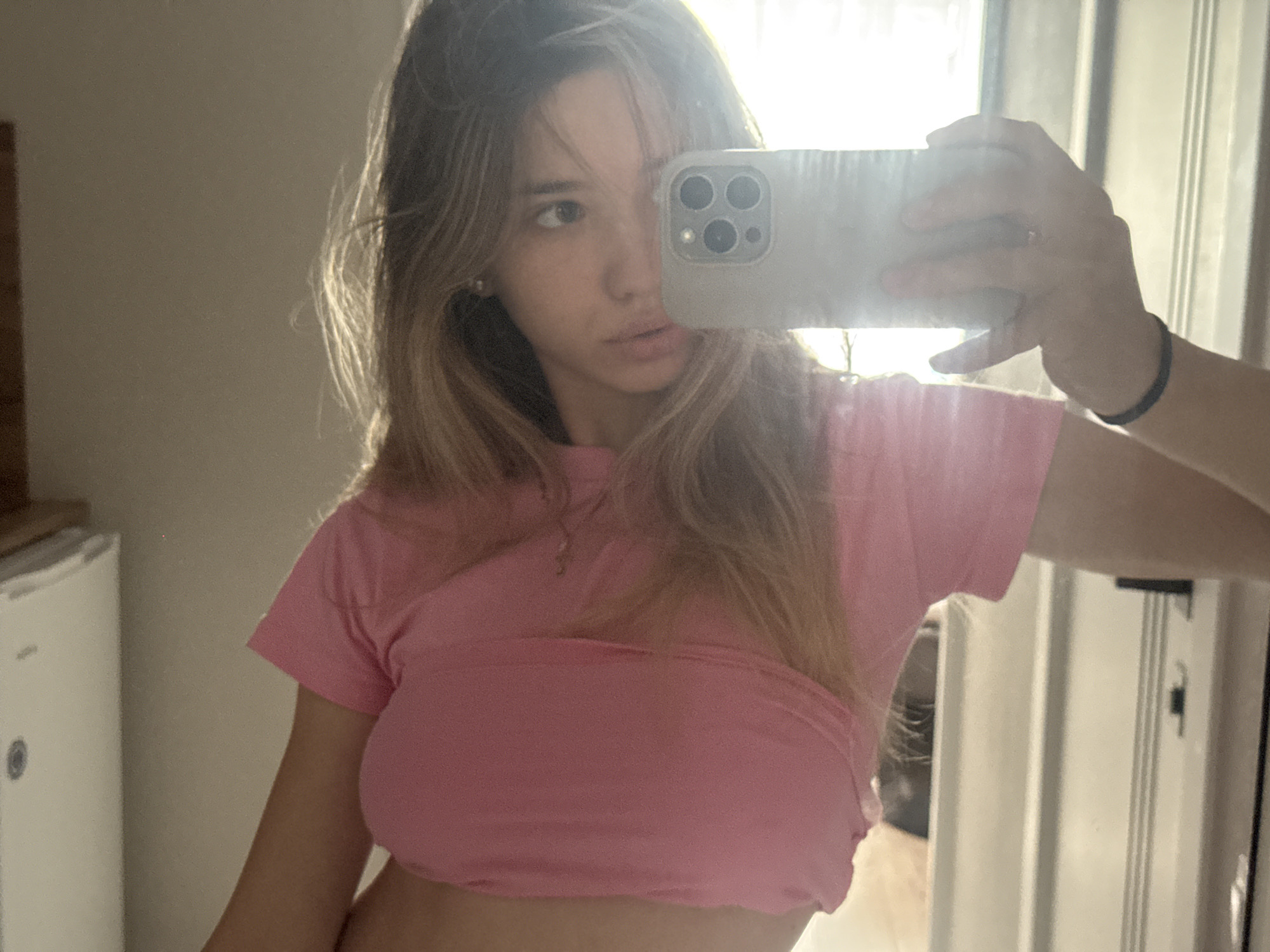A young woman with blonde hair is taking a mirror selfie in what appears to be a room with a white refrigerator on the left and a door with vertical blinds on the right. She is wearing a pink t-shirt and a black bracelet on her right wrist. She is holding a smartphone in her right hand, which is reflecting in the mirror. The lighting suggests it is daytime, possibly indoors near a window. There is no indication of the location being Kazan, Russia.