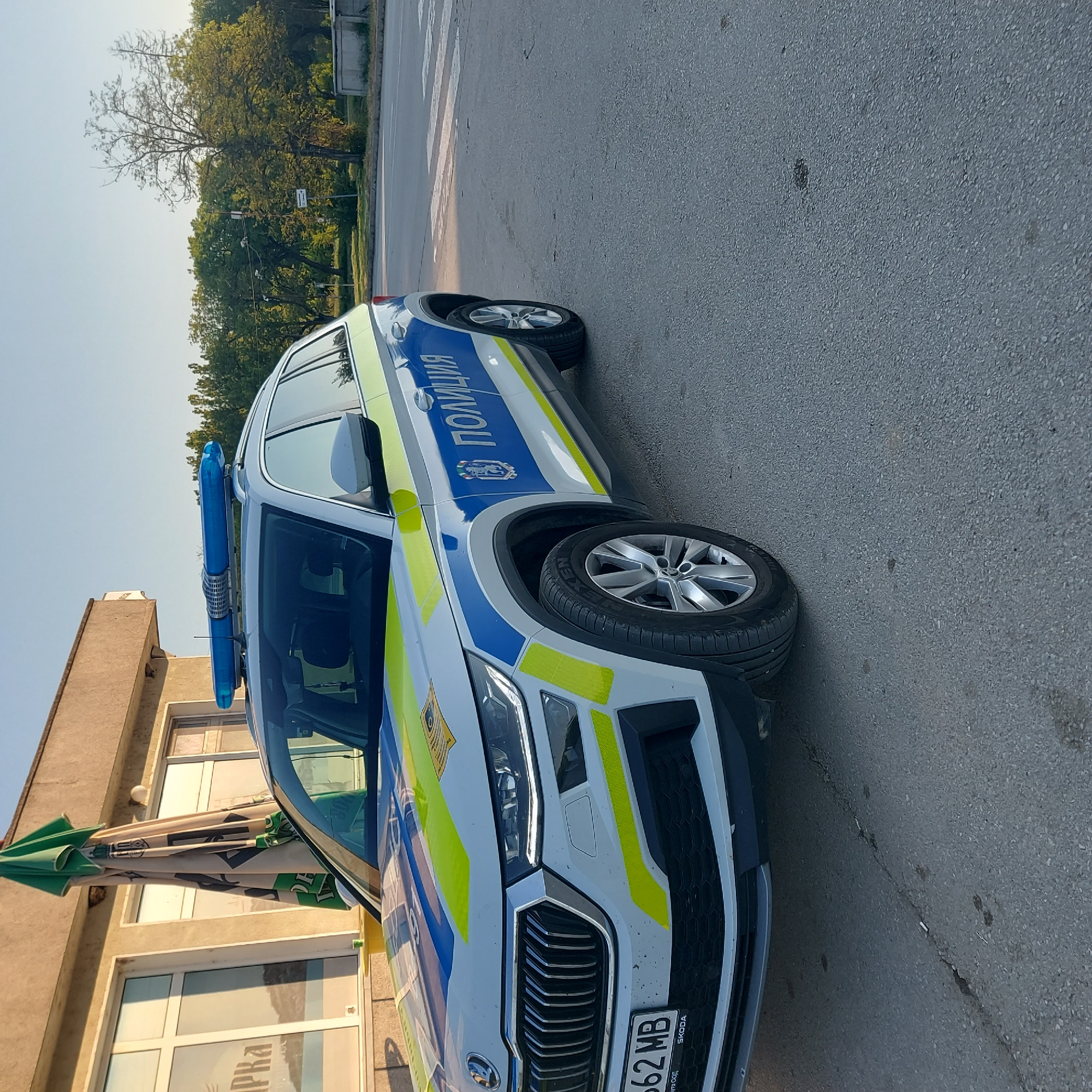 A white and blue Škoda police car is parked on a paved road next to a building with closed storefront windows and a folded green umbrella. The car's license plate reads "PB62MB" and it has blue lights on top. The word "ПОЛИЦИЯ" (Police) is written in Cyrillic script on the car. In the background, there is a tree line and a clear sky. The photograph shows a street setting in Strazhitsa, Bulgaria. There are no visible people.
