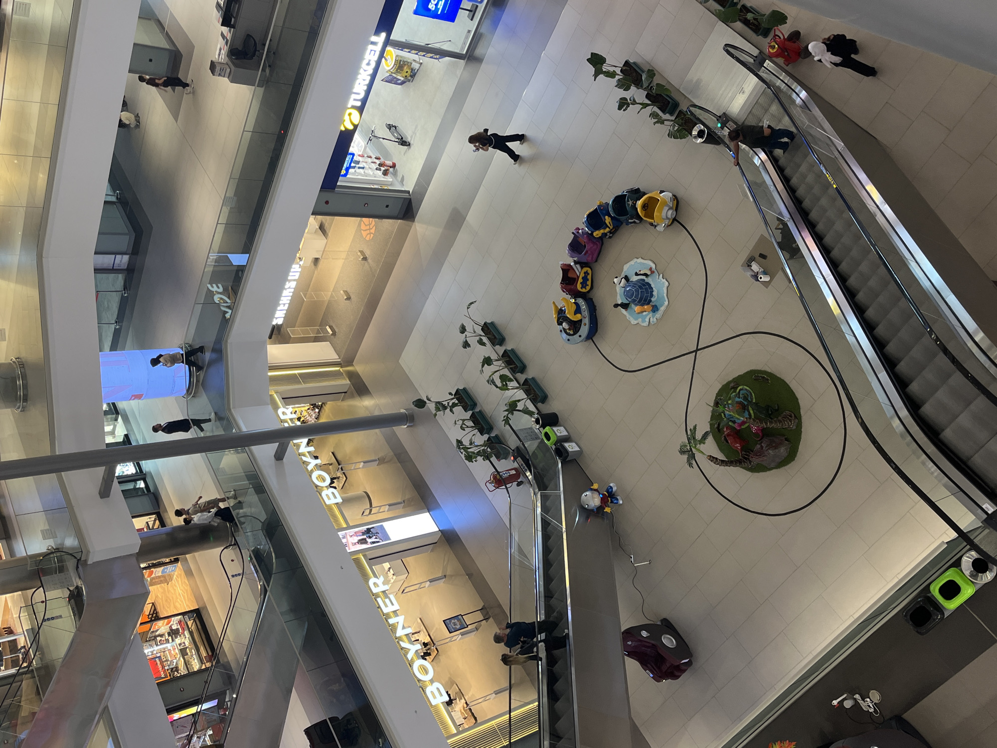 The image depicts the interior of a shopping mall in Alanya, Turkey, viewed from an elevated perspective. The mall features multiple levels with various retail stores, including "Turkcell" and "Boyner," as indicated by signage. People are seen walking on different levels. The central area on the ground floor includes a carousel-style kiddie ride, a small fountain, and a circular decoration featuring artificial plants. Escalators are present, facilitating vertical movement within the mall. Trash receptacles are also visible. The overall atmosphere appears well-lit and modern.