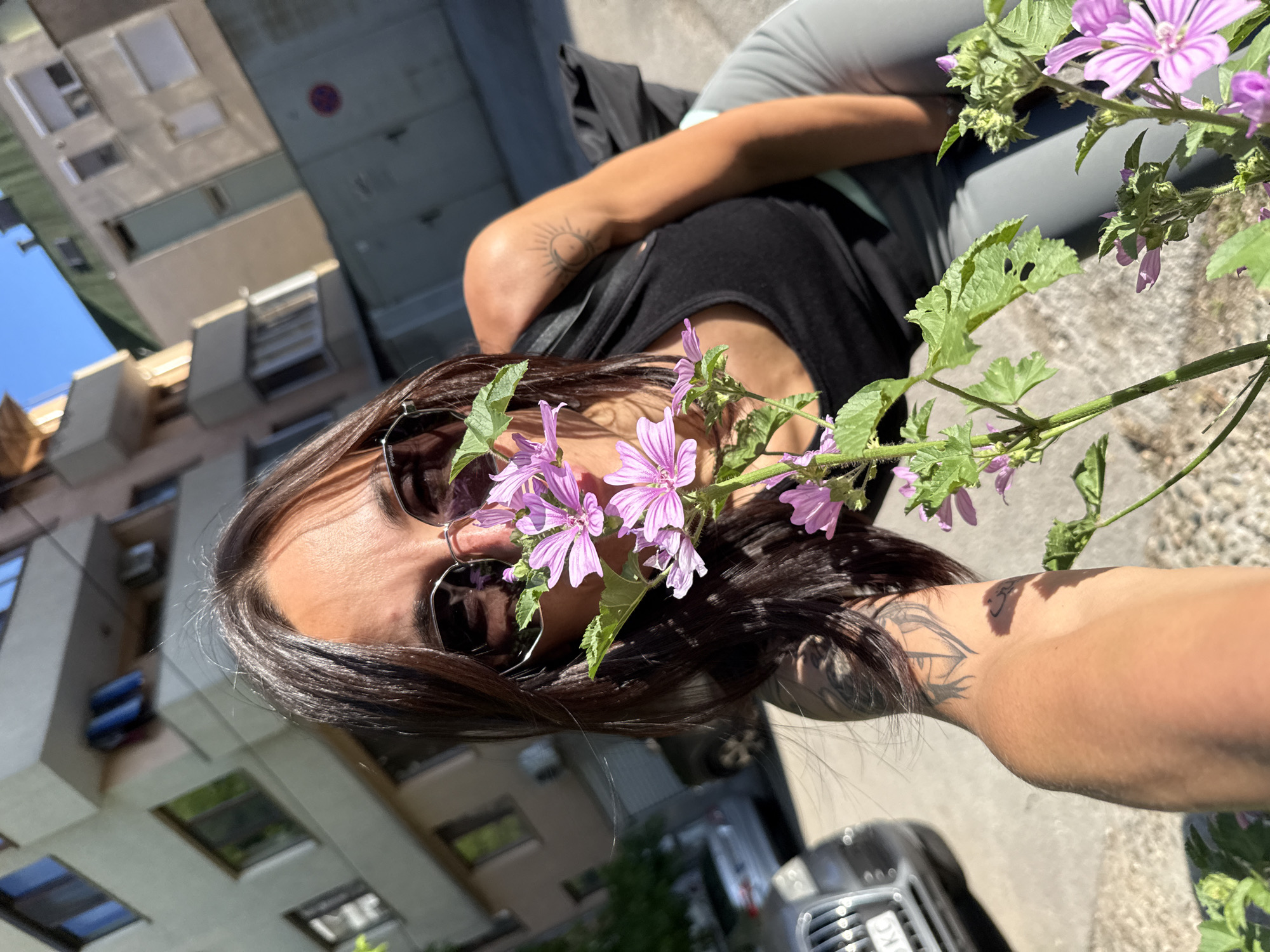 A woman wearing sunglasses and holding a branch of purple flowers is photographed in Pleven, Bulgaria. She has brown hair and tattoos on her arms and neck. She is wearing a black top and grey pants. The background shows buildings, a car, and a street. The image is rotated, making the buildings appear tilted. The location is an urban area in Pleven, Bulgaria, during daytime.
