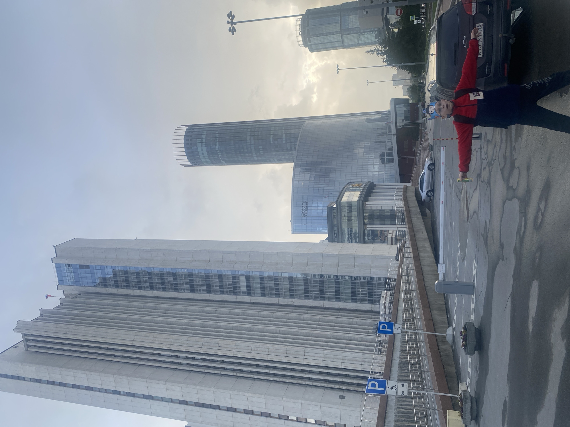 The image depicts a street scene in the **Yekaterinburg-City business district** of Yekaterinburg, Russia, under an overcast sky.
In the foreground, a young male stands with arms outstretched, holding a yellow-green cylindrical object in his left hand. He wears glasses, a blue and red fleece jacket, black pants, and a blue cartoon-themed crown on his head. His mouth is slightly open. Behind him to the right, a dark maroon Chevrolet station wagon (Russian license plate В 973 КУ 196 RUS) is parked. The asphalt ground is wet, featuring white parking markings, two concrete planters with flowers, and a grey parking barrier with a horizontal arm. A concrete ramp with metal railings ascends to the left, marked by two blue 'P' signs for disabled parking, one indicating '5M'.
Dominating the left side of the frame is the **House of Soviets (Government of Sverdlovsk Oblast building)**, a tall, light-colored concrete structure characterized by prominent vertical fins and a blue-white-red flag (Russian flag) flying from its peak. Its facade incorporates reflective glass sections. In the midground, the **Hyatt Regency Ekaterinburg** hotel appears as a tall, cylindrical glass skyscraper with a distinctive spire-like crown. Below it, a curved glass structure with the 'Hyatt Regency' logo is visible. Further in the background on the right, another slender, taller cylindrical skyscraper, identified as the **Iset Tower**, is partially visible. Streetlights and scattered trees are present in the background.