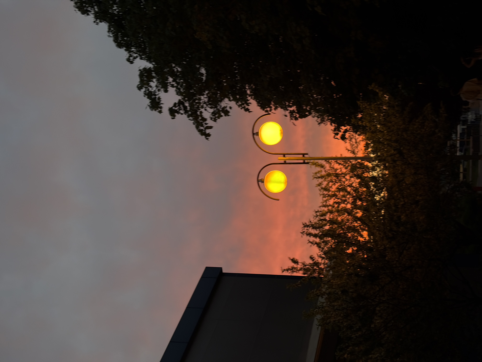 The photograph depicts a street scene in Goryachiy Klyuch, Russia, at dusk. Two globe-shaped streetlights, both illuminated with a warm yellow glow, dominate the center. The sky behind the streetlights is a mix of gray and orange hues, suggesting a sunset or twilight. A building with a dark gray exterior is visible on the lower left, its angular roof contrasting with the softer shapes of the sky and foliage. Trees frame the scene; their branches and leaves appear silhouetted against the sky. In the background, blurred forms suggest a parking lot with parked vehicles.