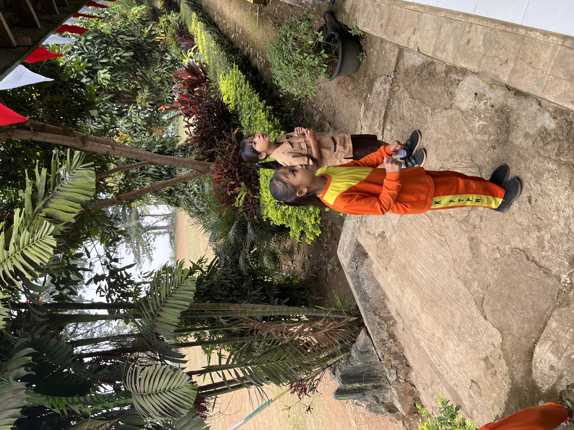 In Gembong, Indonesia, two young girls stand on a concrete pathway lined with lush greenery and potted plants. The girl in the foreground wears an orange long-sleeved shirt with yellow accents and matching orange pants with yellow lettering. The girl behind her is dressed in a tan uniform with a brown skirt. Both girls are looking upwards and to the right, their attention seemingly focused on something outside the frame. In the background, palm trees and other tropical vegetation thrive, suggesting a warm, humid climate. Red and white triangular flags are strung across the top of the frame, hinting at a festive or celebratory occasion.