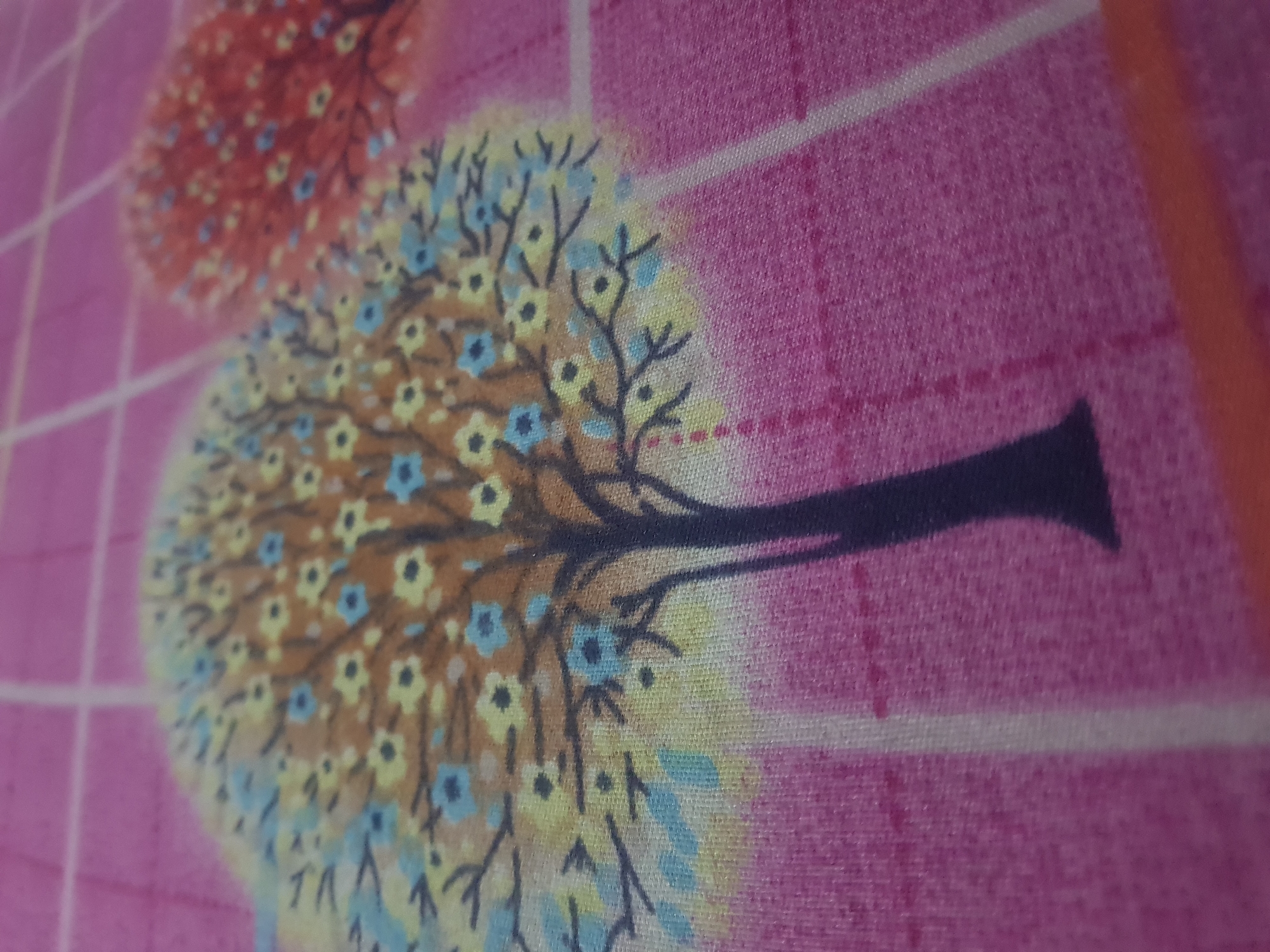 The image is a close-up of a pink fabric with a grid pattern. A stylized tree with a brown trunk and yellow and blue flowers is printed on the fabric. The tree is depicted in a semi-abstract style, with the branches extending outwards and the flowers scattered amongst them. Another tree, with red flowers, is partially visible on the right side of the image. The fabric appears to be a textile, possibly part of a bedsheet or tablecloth. The lighting suggests an indoor environment. The location is Unknown, Unknown.