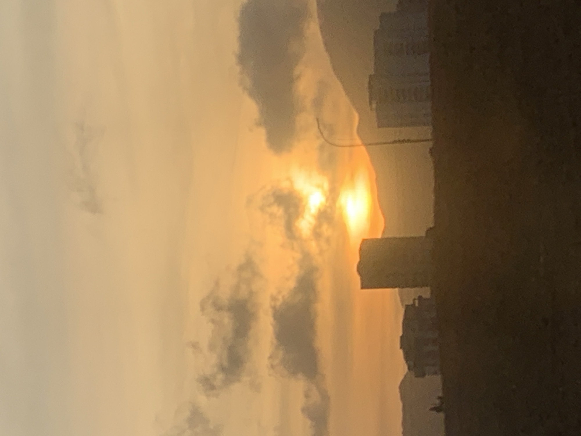 The image depicts a sunset in Porlamar, Venezuela, with buildings silhouetted against the bright, golden light. Clouds are scattered across the sky, partially obscuring the sun. There's a dark land mass on the right side of the frame.