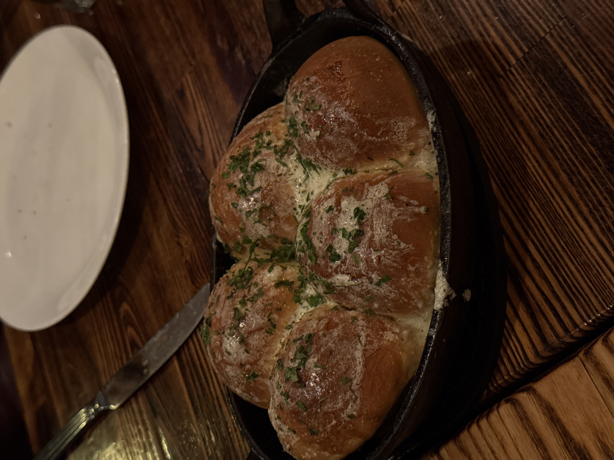 A cast iron skillet filled with six rolls topped with a white substance and green herbs sits on a wooden table. A white plate and knife are also visible on the table. The location is unknown.