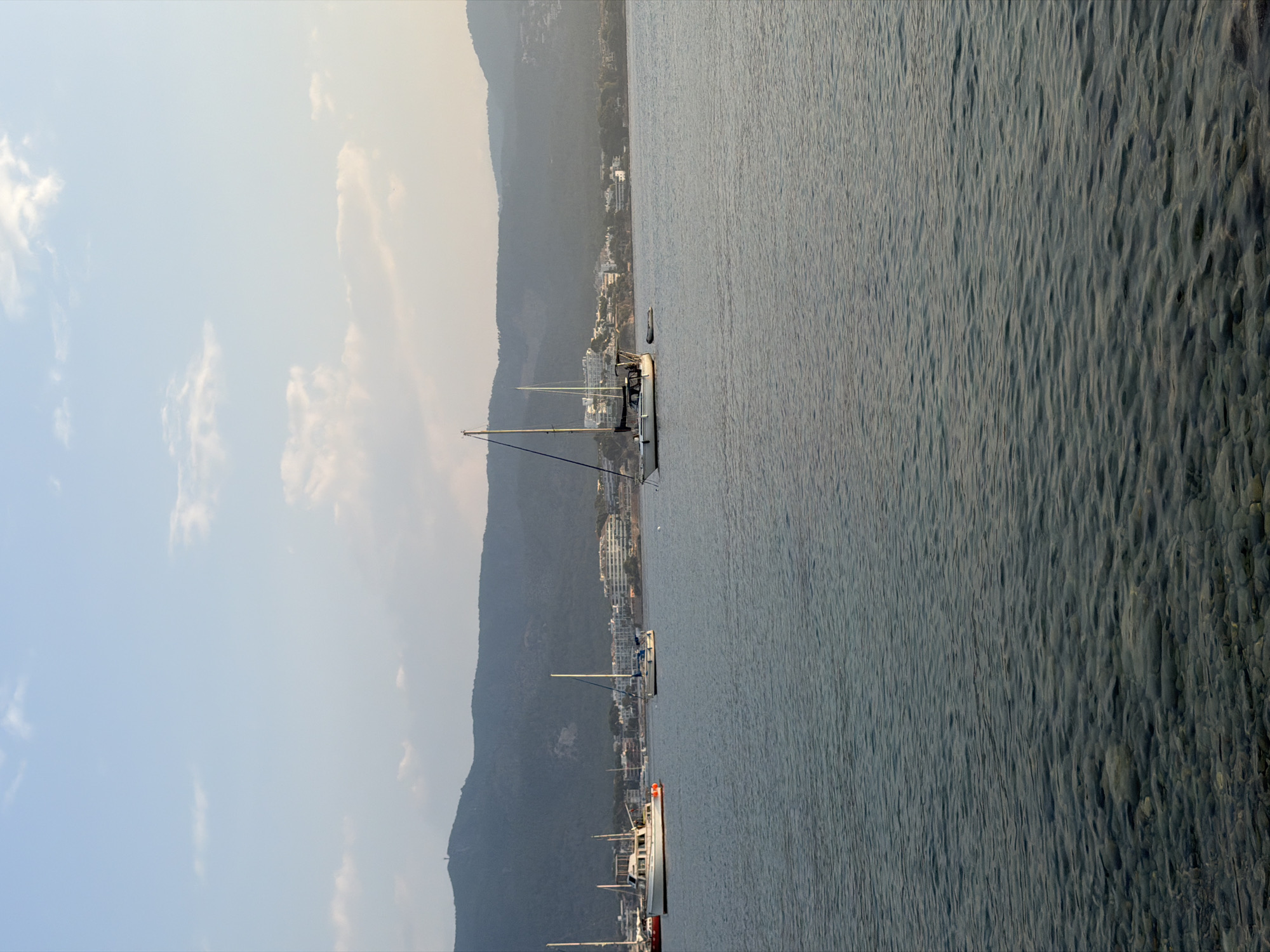 The image depicts a tranquil coastal scene in the municipality of Calvià, Spain. Several sailboats are anchored in the calm, clear turquoise waters of the bay. In the background, a verdant, mountainous landscape rises, dotted with white buildings that suggest a coastal town or resort. The sky above is a pale blue, with wispy white clouds adding texture. The foreground shows the seabed composed of smooth, rounded pebbles, visible through the shallow water. The overall atmosphere is serene, indicative of a peaceful day at sea.