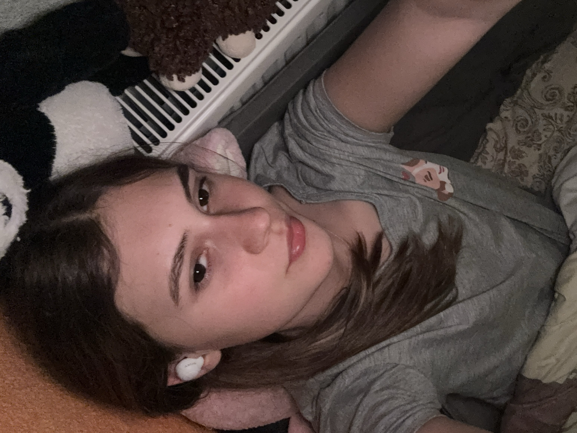 A young woman is captured in a close-up, upward-angle selfie, lying down in an indoor setting in Panagyurishte, Bulgaria. She has medium brown hair, dark eyes, and light skin, looking directly at the camera with a calm, slightly smiling expression. She is wearing a grey t-shirt adorned with a small, light-colored graphic design, possibly a cartoon character's head, on the chest. A white earbud, clearly marked with the brand "SONY," is visible in her right ear, suggesting she is listening to something.
Her head rests on a soft, light pink object, likely a pillow. Around her are various plush items, including a black and white furry toy (possibly a panda) in the upper left, and a brown, textured plush toy resting near a white radiator grill in the upper right. A patterned blanket or pillow is visible on the right side of the frame. The overall lighting is dim, indicating it might be evening or nighttime, enhancing the cozy and relaxed atmosphere of the scene.