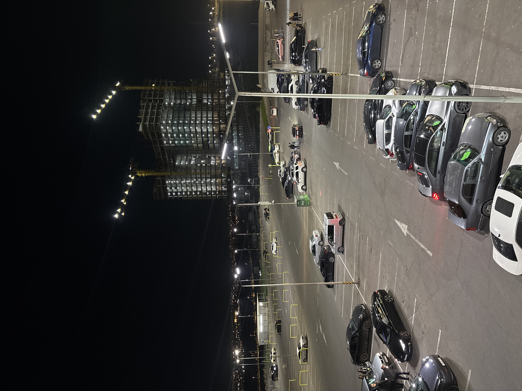 The image depicts a nighttime scene in Yerevan, Armenia. A multi-story building under construction with visible cranes is illuminated to the left. Adjacent to it is a large, well-lit parking lot with numerous parked cars. People are seen standing and interacting near some of the vehicles. Streetlights are spaced throughout the parking area and along the horizon. The parking lot is marked with parking lines and directional arrows.