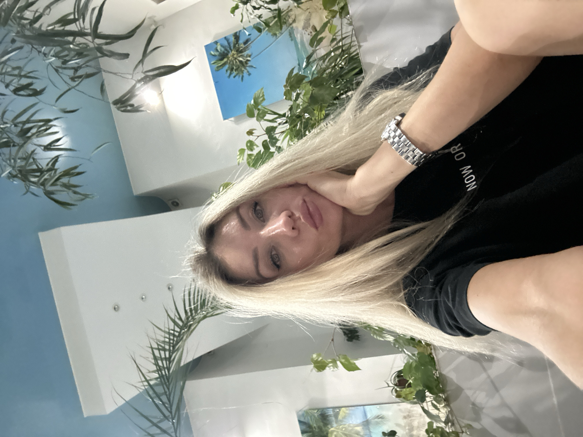 A woman with blonde hair is taking a selfie. She is wearing a black t-shirt with the words "NOW OR" visible on it, and a silver watch on her left wrist. She is resting her chin on her right hand. The background shows a tropical-themed painting of a beach with palm trees, lush green plants, and a light blue ceiling. The overall lighting suggests an indoor environment, possibly a cafe or restaurant. The location is not specified as Armavir, Russia.