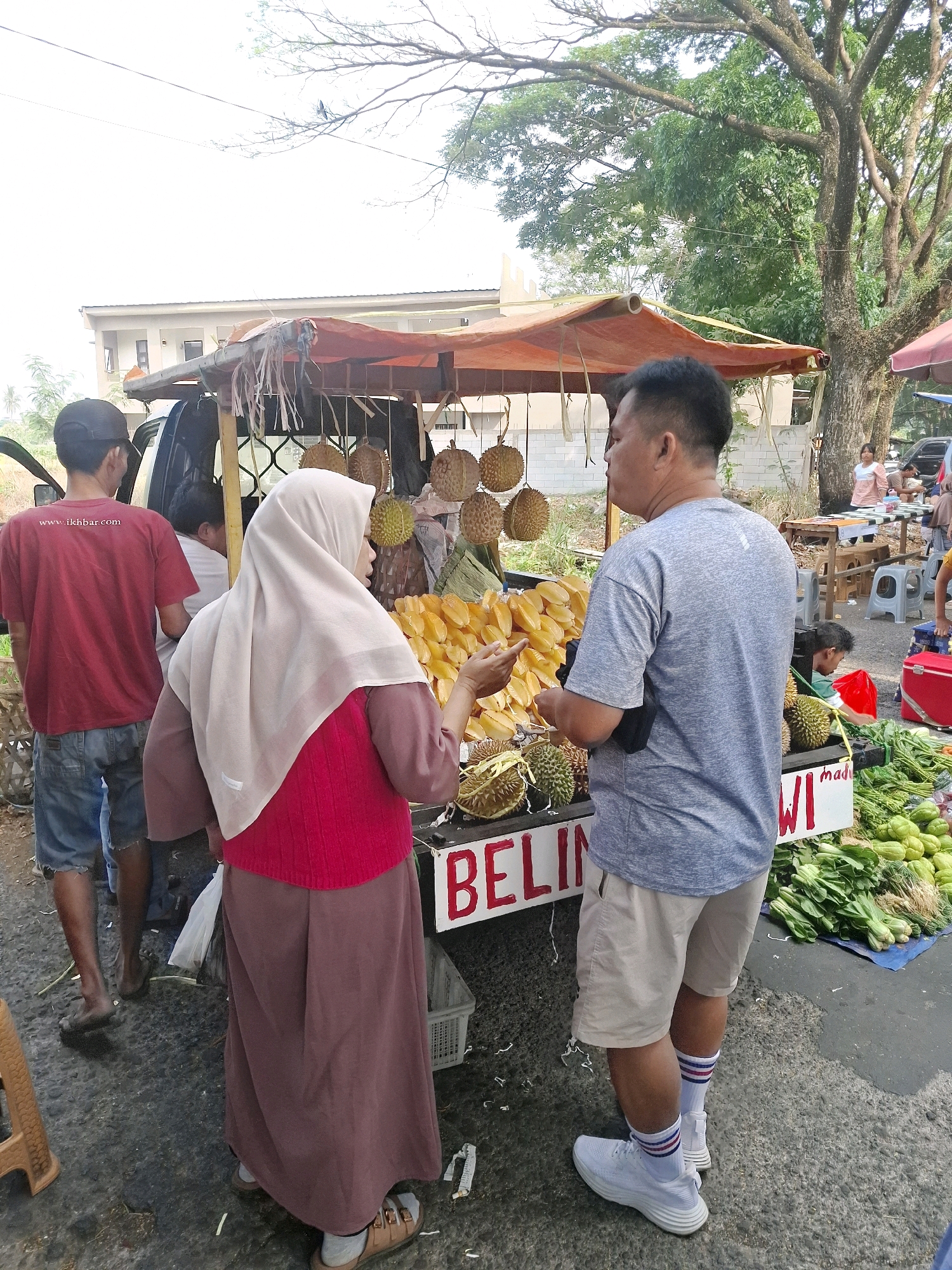 In Karangtengah, Indonesia, an outdoor market scene unfolds under a bright sky. A temporary stall, adorned with an orange tarp, displays a variety of fresh produce. Durian fruits hang from the stall, while piles of starfruit and bundles of green vegetables are arranged for sale. A man in a grey t-shirt and beige shorts engages with a woman wearing a light beige hijab and a bright red vest over a maroon skirt. The woman is pointing towards the starfruit. Behind them, other individuals are visible, including a man in a red t-shirt and blue jeans standing near a vehicle. The ground is a mixture of asphalt and dirt, with scattered debris. The environment suggests a lively marketplace, with trees and buildings in the background.