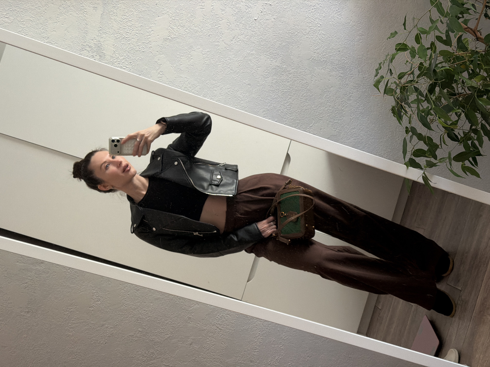 A female is taking a selfie in a mirror. She is wearing a black top, a black leather jacket, brown pants, and black shoes. She is holding a green purse. The background is a wall and a plant. The location is Perm, Russia.