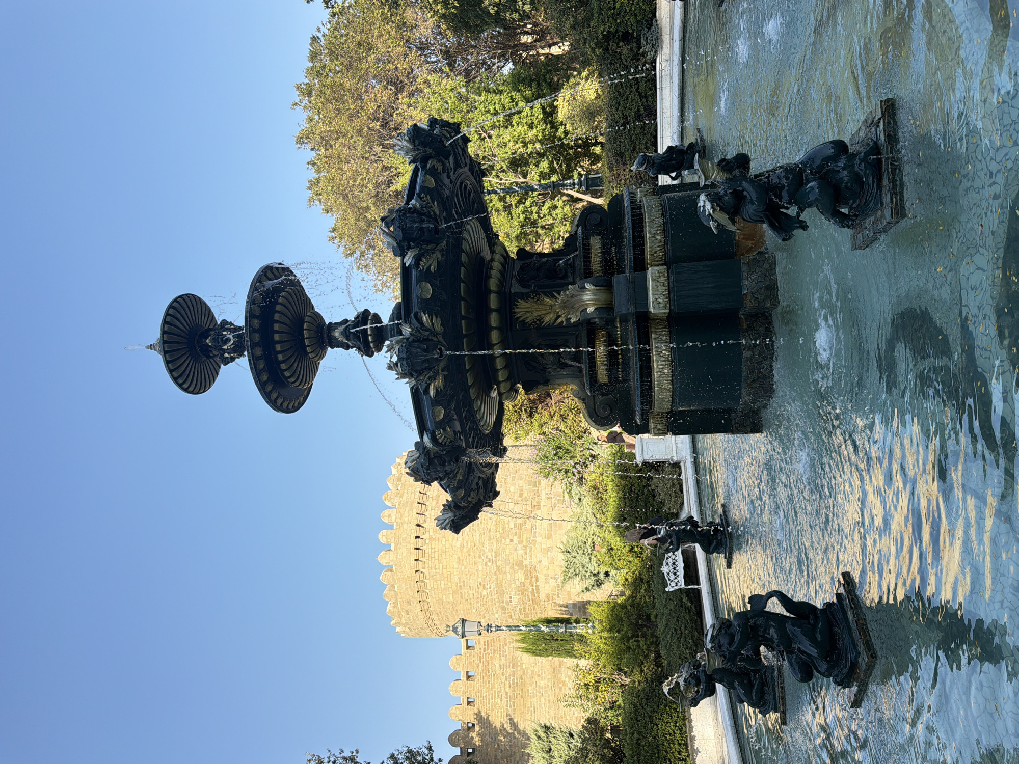 The image depicts a public fountain in Sabail Raion, Azerbaijan, with a historical stone wall in the background. The fountain is a multi-tiered, dark-colored structure adorned with decorative elements. Water cascades from the upper tiers, splashing into the pool below. Around the central fountain, several small, dark sculptures of figures are partially submerged in the water, each situated on a stone base. One sculpture in the foreground features a figure holding what appears to be a torch. In the middle ground, a white park bench is visible, with a person standing near it. A woman with dark hair is also visible standing in the background, closer to the fountain. The surrounding area is landscaped with green bushes and trees. The sky is clear and blue, suggesting a daytime setting. The stone wall in the background is cylindrical with crenellated battlements, indicative of ancient architecture. A lamppost stands to the left of the wall. The overall scene conveys a serene and historical ambiance within an urban park setting.
