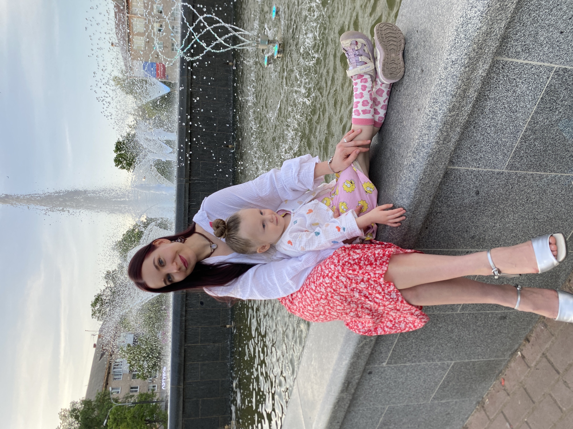 A woman and two children are photographed at a fountain in Chaikovsky, Russia. The woman, with long burgundy hair, is seated, wearing a white blouse and a red and white floral print skirt, and silver sandals. A young child with their hair in a bun reclines on her lap, wearing pink pajamas with yellow cartoon character prints and a white shirt with heart prints. A second child with pink and white patterned leggings and purple sandals sits next to them. The fountain behind them has multiple water jets, and buildings are visible in the background.