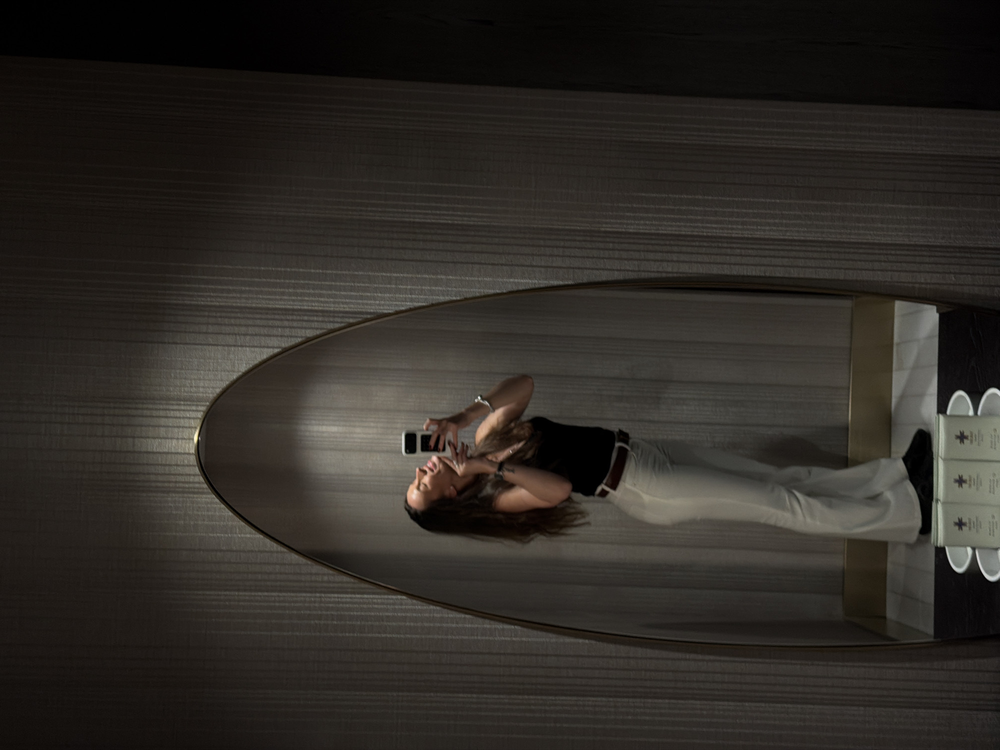 A woman is reflected in an oval mirror, taking a selfie with a smartphone. She is wearing a black top, white pants with a brown belt, and black shoes. She has long brown hair. The mirror is mounted on a textured wall with horizontal lines. To the right of the mirror, on a shelf, there are three white cups and three small boxes with a purple logo. The location is Budva, Montenegro.