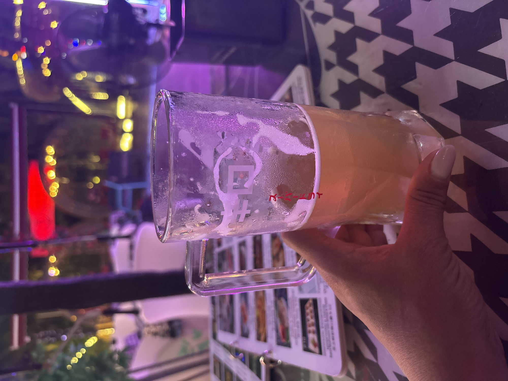 A hand holds a partially full glass mug of beer, condensation visible on the glass, placed on a table with a black and white star pattern. In the background, the scene is blurred with colorful lights suggesting an outdoor or restaurant setting at night, possibly in Jiyang District, China. A menu with food images is visible to the left of the mug.