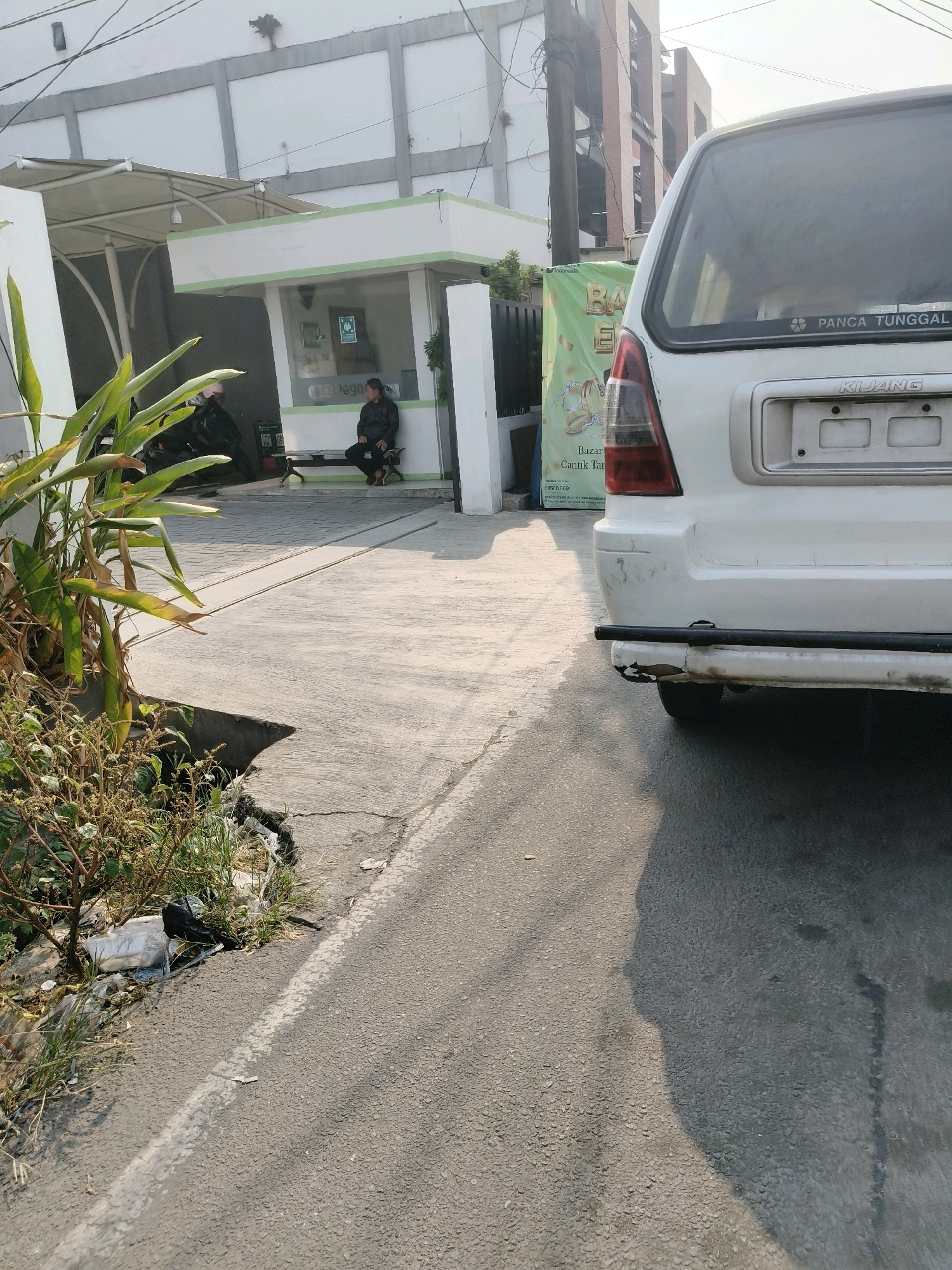 A white van is parked on the right side of the frame, facing away from the viewer, with its rear to the camera. The van has a sticker on its rear window that reads "PANCA TUNGGAL" and the model name "KIJANG" is visible above the license plate area. The van appears to be parked on an asphalt surface.
To the left of the van, there is a small, open-air structure with a white roof supported by metal poles and a white facade with green trim. Inside this structure, a person wearing a dark jacket and pants is seated on a bench, facing towards the left side of the frame. Next to the person, there is a motorcycle parked under a covered area.
In the foreground on the left, a plant with large, green leaves and some dry, brown foliage is visible, growing out of a cracked concrete area. This area also has some scattered trash, including plastic bags and bottles. The ground is a mixture of concrete and asphalt, with a white line painted on the asphalt. In the background, there is a building with a white facade and a series of horizontal grey bands. There are also some electrical wires and a utility pole visible.
The image appears to have been taken in South Tangerang, Indonesia, possibly near a business or residential area, given the presence of the van, the small structure that could be a security post or a small shop, and the surrounding buildings. The lighting suggests it is daytime, with sunlight casting shadows.