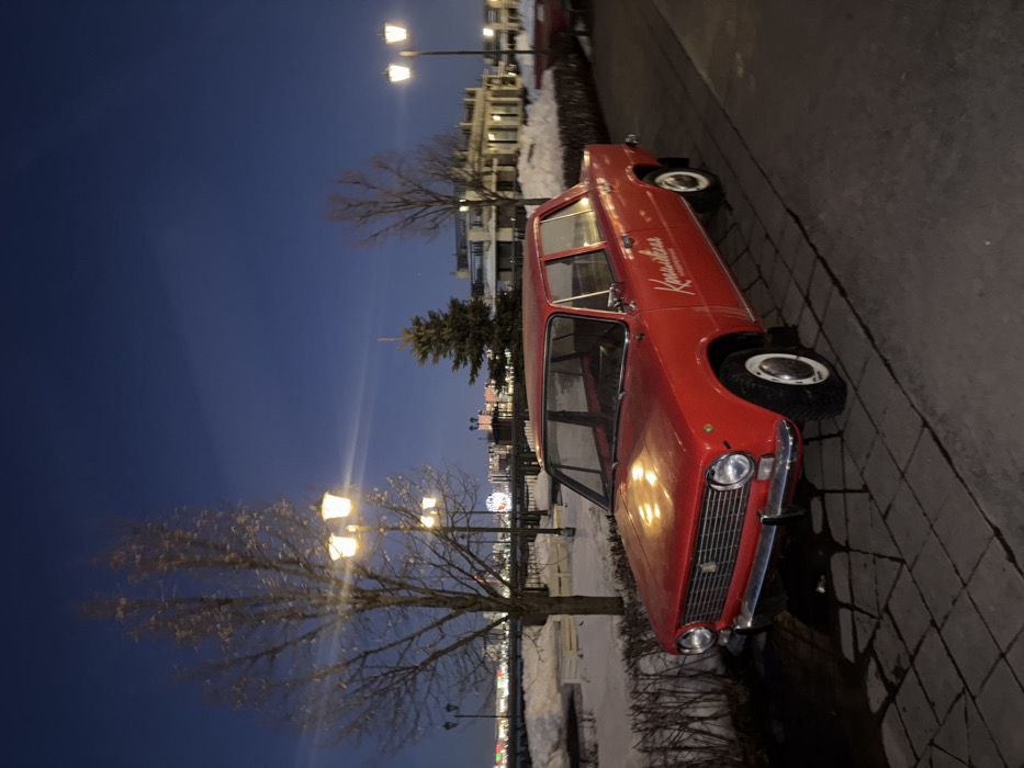 A red VAZ-2101 (Lada Zhiguli) four-door sedan is prominently displayed in the foreground, angled slightly to the left, on a dark gray paved surface composed of rectangular stone blocks. The vehicle is bright red with chrome bumpers, round headlights, and white hubcaps. White Cyrillic lettering, "Канава," is present on the front passenger door.
The scene is set at dusk or nighttime, under a deep blue sky, illuminated by multiple classic-style streetlights with warm yellow light, casting reflections on the car's hood and body. Directly behind the car on the left is a leafless deciduous tree. Further right, another leafless tree and a coniferous tree are visible. A low, dark hedge runs parallel to the paved area, separating it from a snow-covered bank.
In the mid-ground, beyond the snow, a body of water is implied, followed by the illuminated skyline of Kazan, Russia. Distinctive features in the background include a prominent Ferris wheel on the left and modern multi-story buildings with structured facades. The presence of snow confirms a winter environment. No individuals are present in the image. The car is stationary. This location is consistent with an urban embankment or promenade area along the Kazanka River or Lower Kaban Lake in Kazan.