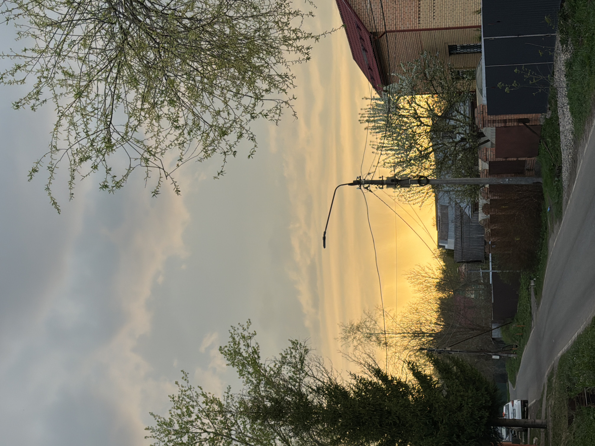 A residential street in an unidentified location. A two-story brick building with a red roof is visible. A light post stands adjacent to the building, with wires extending from it. Trees with budding leaves frame the sky. A paved road runs alongside the buildings. A vehicle is partially visible at the bottom of the image. The sky is a mixture of gray clouds and a yellow hue.