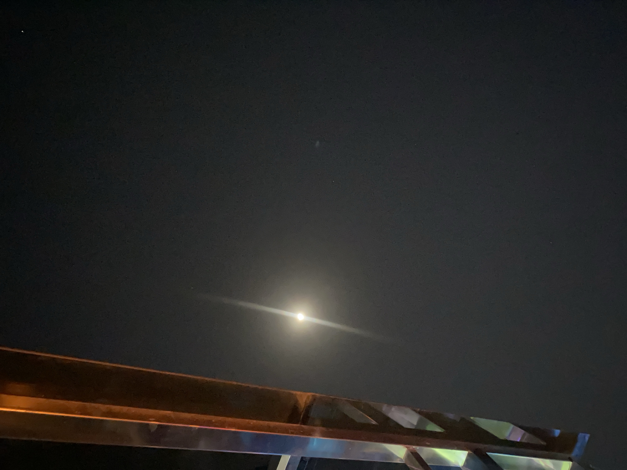 The image depicts a night scene in Sabon Gari West, Nigeria, dominated by a dark sky. A bright, luminous full moon is positioned slightly left of the center of the frame, casting a prominent glare with visible light rays radiating outward. In the foreground, a section of an orange-colored metallic structure with reflective surfaces is visible on the left side, angled towards the sky. The structure appears to be part of a larger architectural element, possibly a balcony railing or a decorative support. A faint, smaller speck of light, potentially a star or a distant light source, is present in the upper right portion of the sky. The overall environment suggests an urban setting at night, with the moon and the artificial structure being the primary focal points.