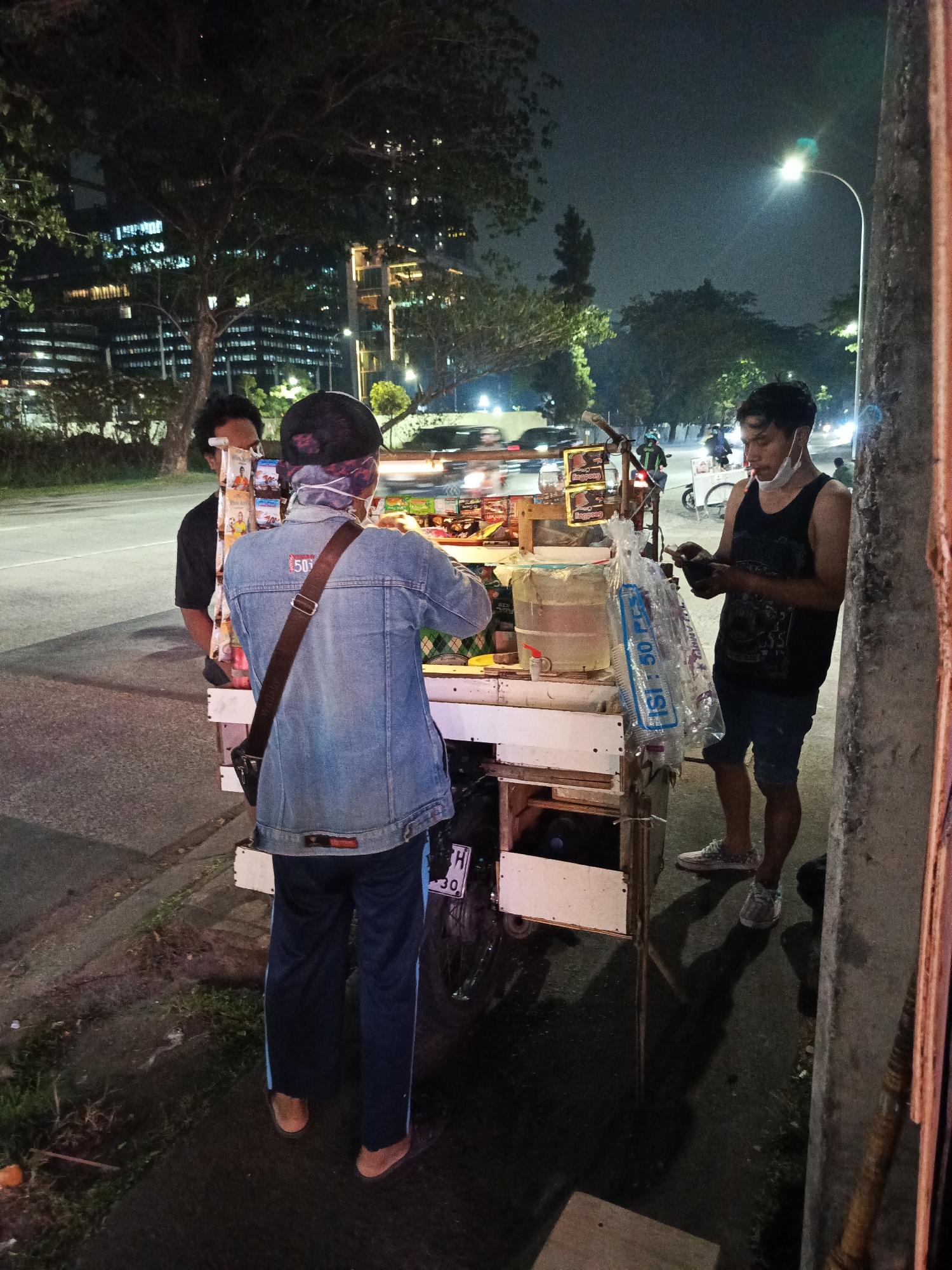 The image depicts a street food cart at night in the city of Tangerang, Indonesia. Two men and one person wearing a denim jacket are visible. The person in the denim jacket, facing away from the camera, is interacting with the food cart. The man in the black tank top, on the right, is holding a mobile phone and wearing a face mask. The second man, wearing a black shirt, is partially visible on the left. The food cart is laden with various items, including packaged snacks, a large water dispenser, and cups. The background shows trees, a street with passing vehicles, streetlights, and illuminated buildings, indicative of an urban environment at night. The scene suggests a commercial interaction taking place on a sidewalk next to a road.