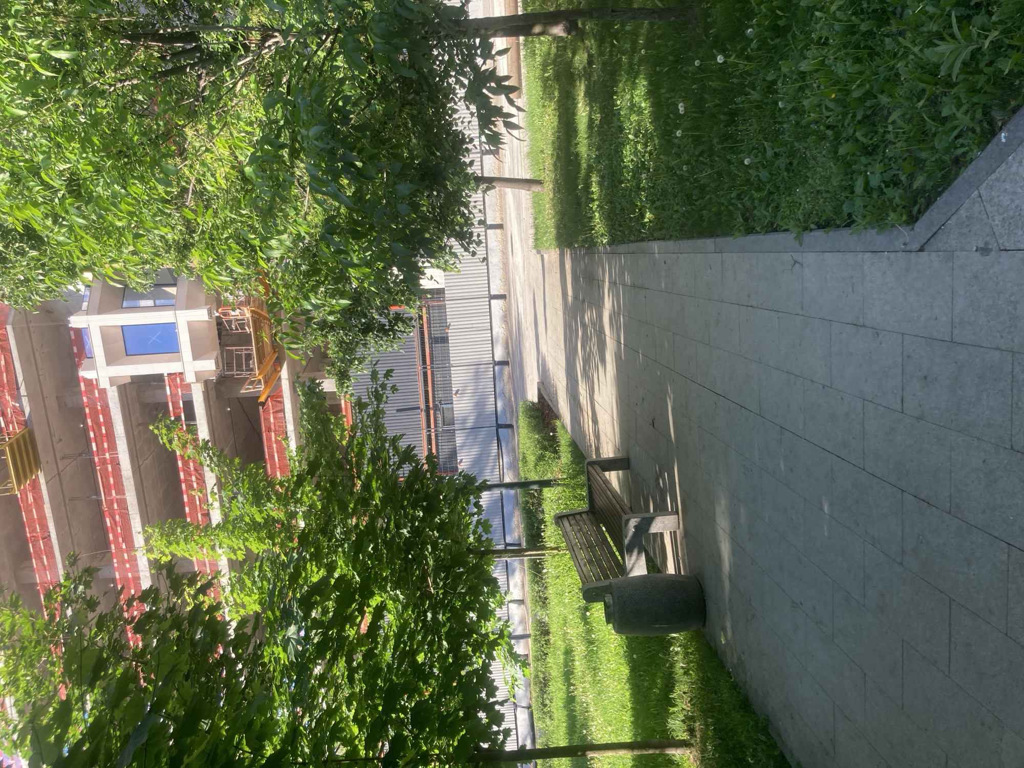 The image depicts a park setting in Moscow, Russia, adjacent to an ongoing construction site. A paved walkway stretches through the frame, bordered by grass and trees. A bench with a concrete base is situated alongside the path. In the background, a building under construction is visible, featuring red safety netting and scaffolding. A temporary corrugated metal fence separates the park from the construction area. The scene is bathed in sunlight, casting shadows on the walkway and grass. There are no people visible in the image.