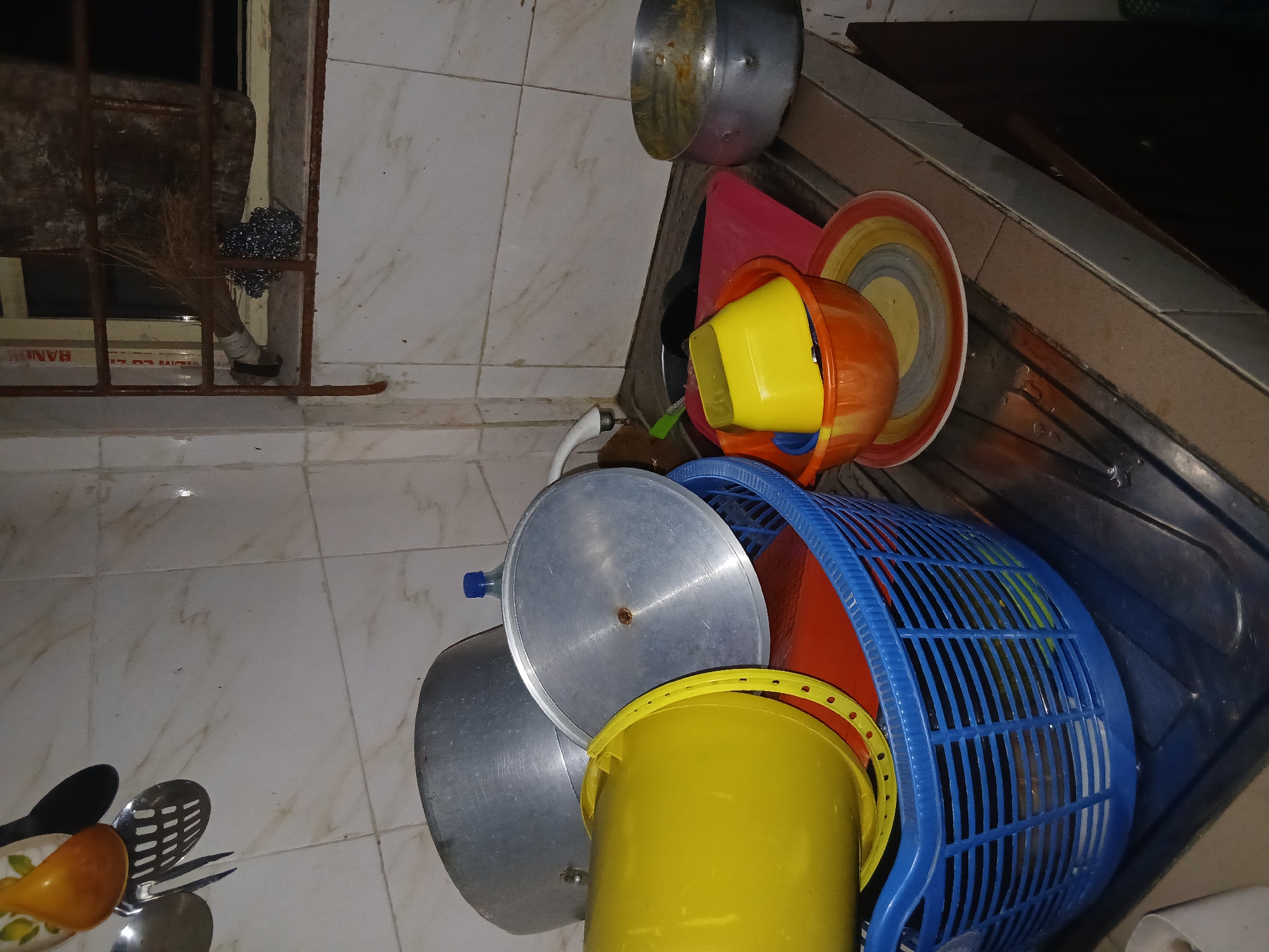 The image shows a cluttered scene in what appears to be a kitchen or washing area. The floor is covered in light-colored tiles with a marble-like pattern. In the foreground, there are several kitchen utensils, including spoons and a slotted spatula, lying on the floor.
The main focus of the image is a collection of various household items. There are several metal pots, a yellow bucket, a yellow funnel inside an orange bucket, a red dishpan, and a blue plastic basket. These items are stacked and overlapping, creating a disorganized pile.
In the background, there is a window with a metal grate and a broom leaning against the wall. Part of a countertop is visible above the utensils.
There is no indication of a specific city or region. The overall impression is one of untidiness and lack of organization.
