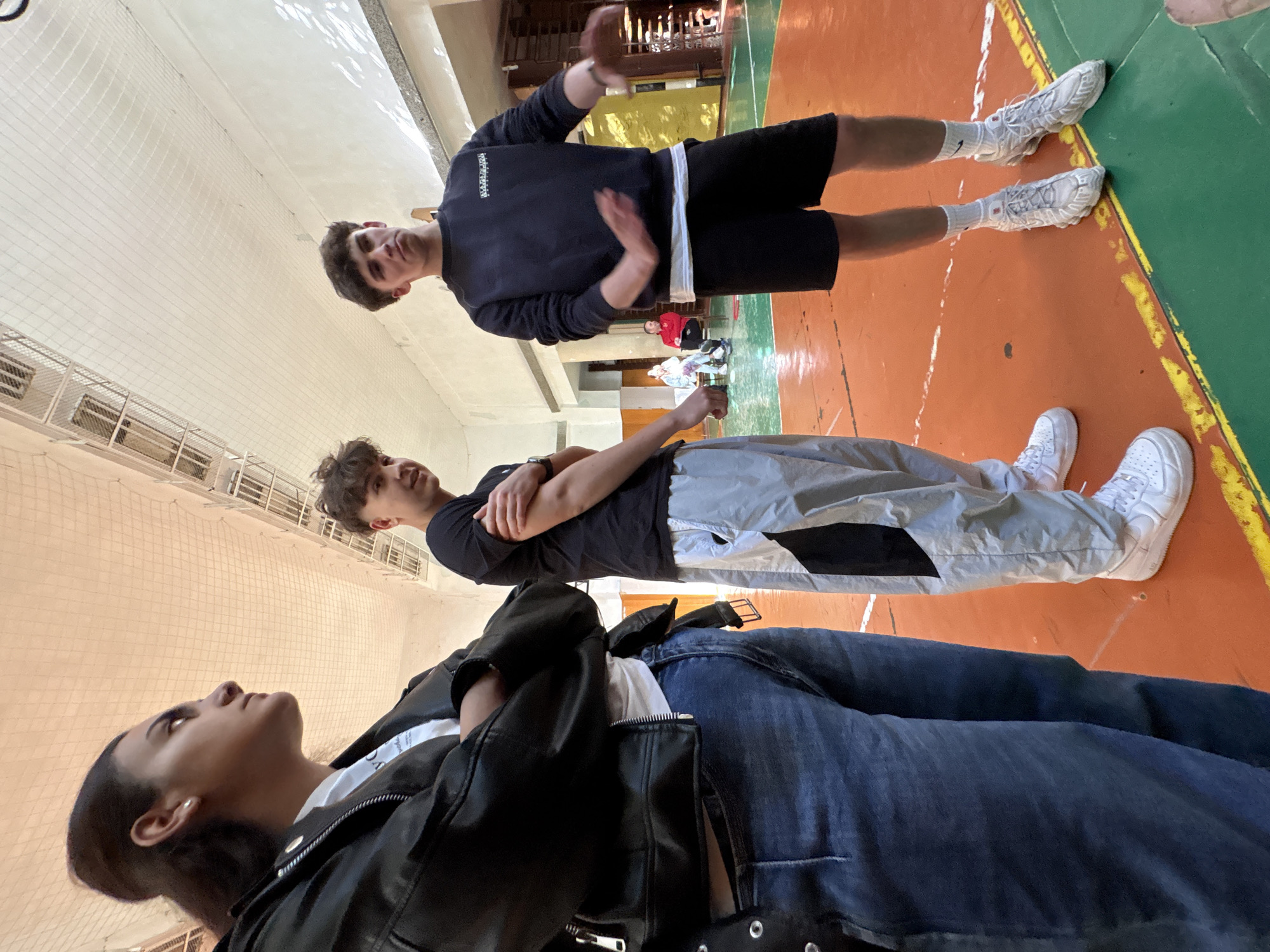 The image shows three people in a gym-like setting, likely in Burgas, Bulgaria, although there is no specific detail to verify. The gym floor is orange with some green areas. The person nearest the camera is a woman wearing a black leather jacket over a white t-shirt and blue jeans, looking upwards. Above her is a man with crossed arms, wearing a black t-shirt and grey/white pants. At the top of the frame is another man wearing a black sweatshirt with white lettering and black shorts, gesturing with his hands. Other people are visible in the background, some sitting.