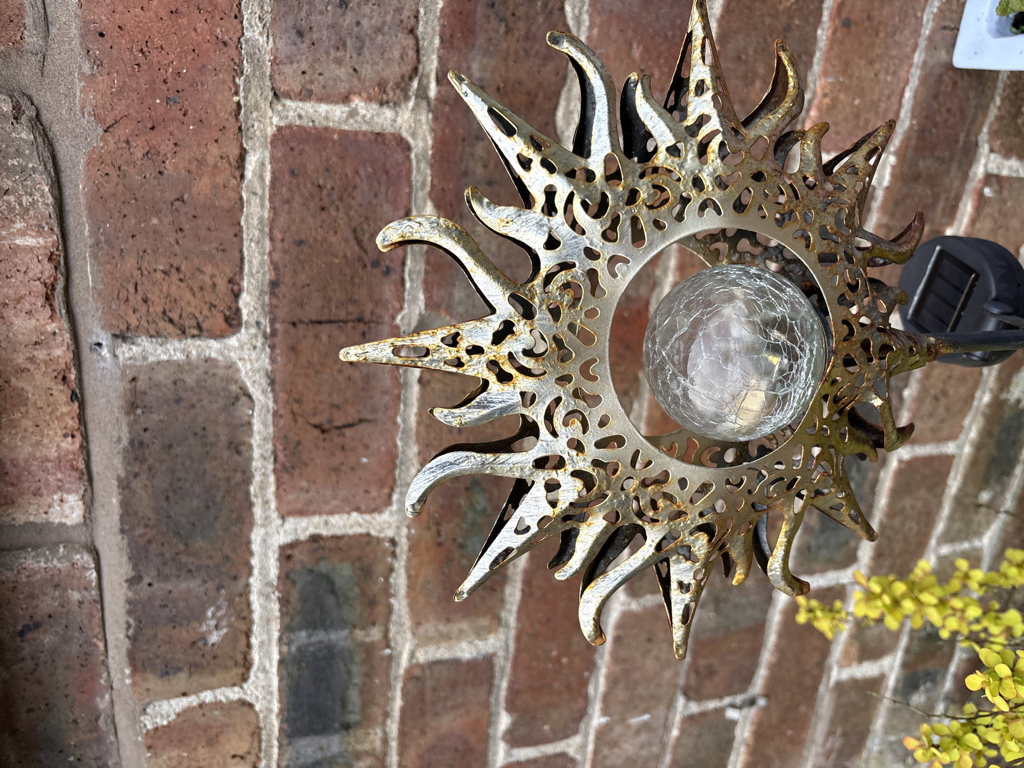 A close-up shot shows a metal sun-shaped garden light fixture with a crackled glass sphere in the center, positioned in front of a brick wall. The metal is colored gold, silver, and a hint of rust. The fixture is viewed at a slight angle, with the solar panel attachment visible to the right. In the lower right, a sprig of small, yellow-leaved foliage appears. The brick wall provides a textured background, featuring traditional red bricks with light gray mortar.