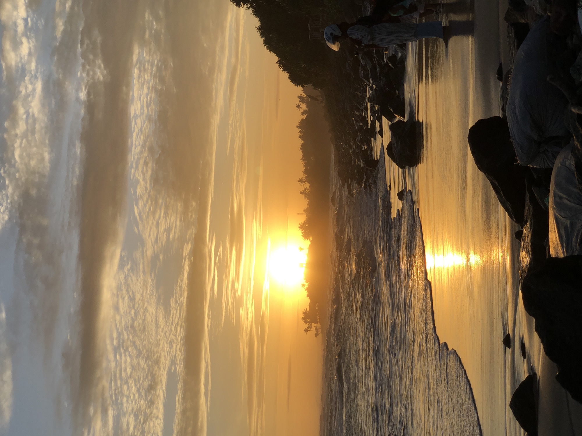 The image depicts a sunset over a rocky beach in Kathaluwa, Sri Lanka. The sun, a brilliant golden orb, is positioned low on the horizon, slightly left of the image center, partially obscured by distant silhouetted tropical vegetation and structures. The sky exhibits a gradient of warm golden-orange tones near the horizon, transitioning to lighter blues higher up, filled with scattered altocumulus and stratocumulus clouds illuminated from below.
The foreground features a wet sandy beach reflecting the intense golden light of the setting sun, creating a luminous path from the water's edge towards the viewer. Dark, choppy ocean waves break on the shore, reflecting some of the overhead light. Numerous dark rocks are scattered along the coastline, both on the sand and partially submerged in the shallow water.
On the right side of the frame, a person stands on the rocky beach, mostly facing away from the camera, observing the sunset. They are wearing a light-colored, possibly striped, long garment over blue pants and a white helmet with a clear visor. A smaller figure is partially visible behind them. To their right, the coastline is densely covered with dark tropical foliage, forming a silhouetted landmass against the sky. In the immediate lower foreground, several dark rocks are visible, along with two light-colored, bagged items, possibly debris, resting on the beach.