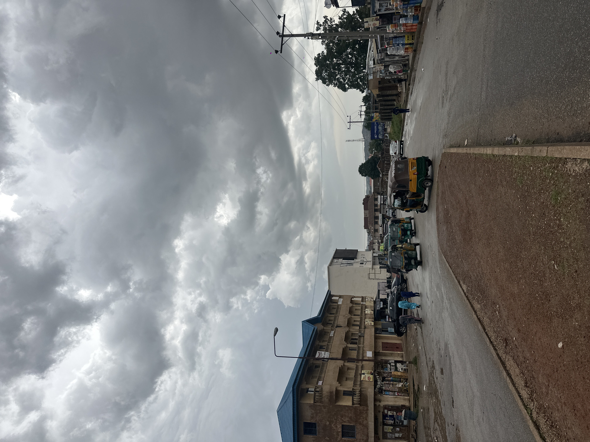 The image depicts a street scene in Jos, Nigeria, under a heavily overcast sky. The day appears to be either early morning or late afternoon, given the diffused, gray light and the absence of harsh shadows. The weather is cloudy, with large, dramatic cloud formations dominating the sky, suggesting an impending or recent rain shower, as the road surface is wet in places.
The scene is a street lined with buildings on the left, housing various shops and businesses, evident from the shelves stacked with goods visible through openings. The buildings are of a modest architectural style, with some showing signs of age. On the right side of the street, there is a paved road and a dirt shoulder.
The street is populated with several auto-rickshaws, commonly known as "Keke Napep" in Nigeria, suggesting active local transportation. Some of these are parked, while others appear to be in motion or waiting for passengers. A few individuals are visible, either walking or standing near the vehicles and buildings, dressed in casual attire. Their postures don't convey strong emotions, but the overall atmosphere is one of everyday activity despite the gloomy weather.
Prominent objects include utility poles with numerous wires crisscrossing the sky, typical of urban infrastructure in many parts of Nigeria. A street lamp stands near a building. A few vehicles, including cars and more Keke Napep, are lined up on the road.
There are no easily discernible signs with readable text, though some posters or advertisements are attached to the buildings. The overall impression is a glimpse into the daily life and streetscape of a bustling area in Jos, Nigeria, characterized by its distinctive transportation, architecture, and the omnipresent cloudy sky.