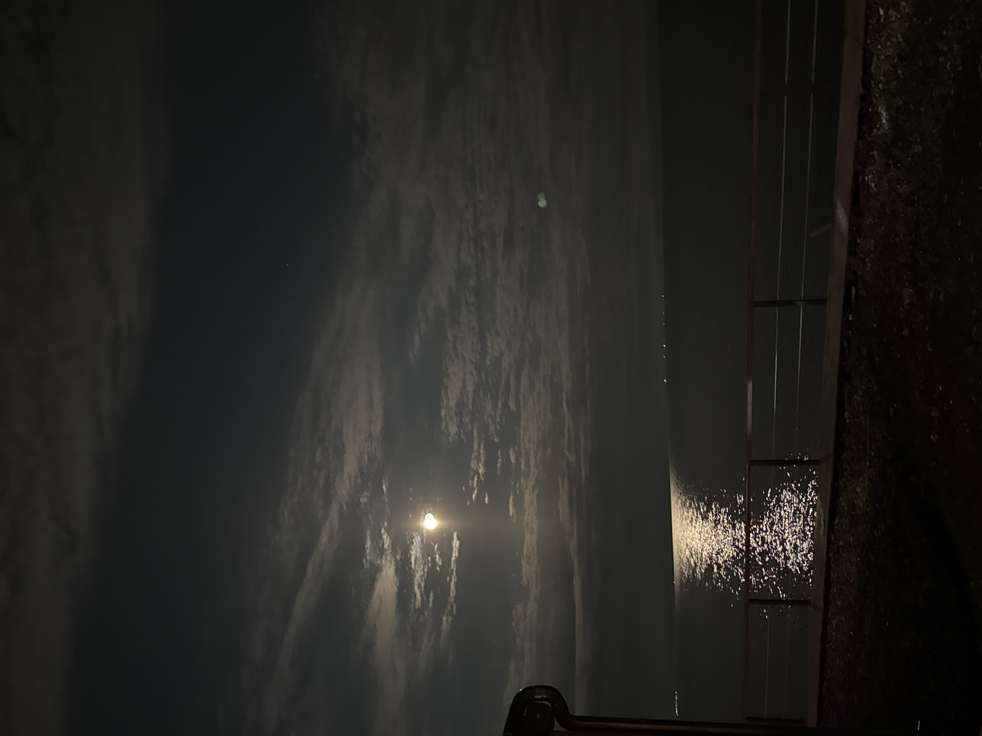 The image captures a serene night scene featuring a bright moon, clouds, and a body of water. The dominant feature is a large, luminous moon, appearing nearly full, positioned in the upper-left quadrant of the frame (when viewed in landscape orientation, rotated 90 degrees counter-clockwise from the original). Its light pierces through scattered, wispy clouds that stretch across the dark night sky, creating a dramatic, textured ceiling.
Below the sky, a reflective body of water shimmers with the moon's light, forming a distinct, sparkling path across its surface, suggesting gentle ripples or movement. In the upper-right portion of the dark sky, a faint, small, light object with a slight green tint is visible, possibly a distant celestial body or a lens flare.
On the right side of the image, dark, linear elements resembling railings or supports of a structure, possibly a dock, pier, or boat, frame the scene. The immediate foreground on this side consists of a dark, textured surface, possibly land or part of a structure, indistinct due to the low light.
The time of day is clearly night, illuminated almost entirely by the moon. The overall atmosphere is quiet and peaceful. There is no visible human activity or text.