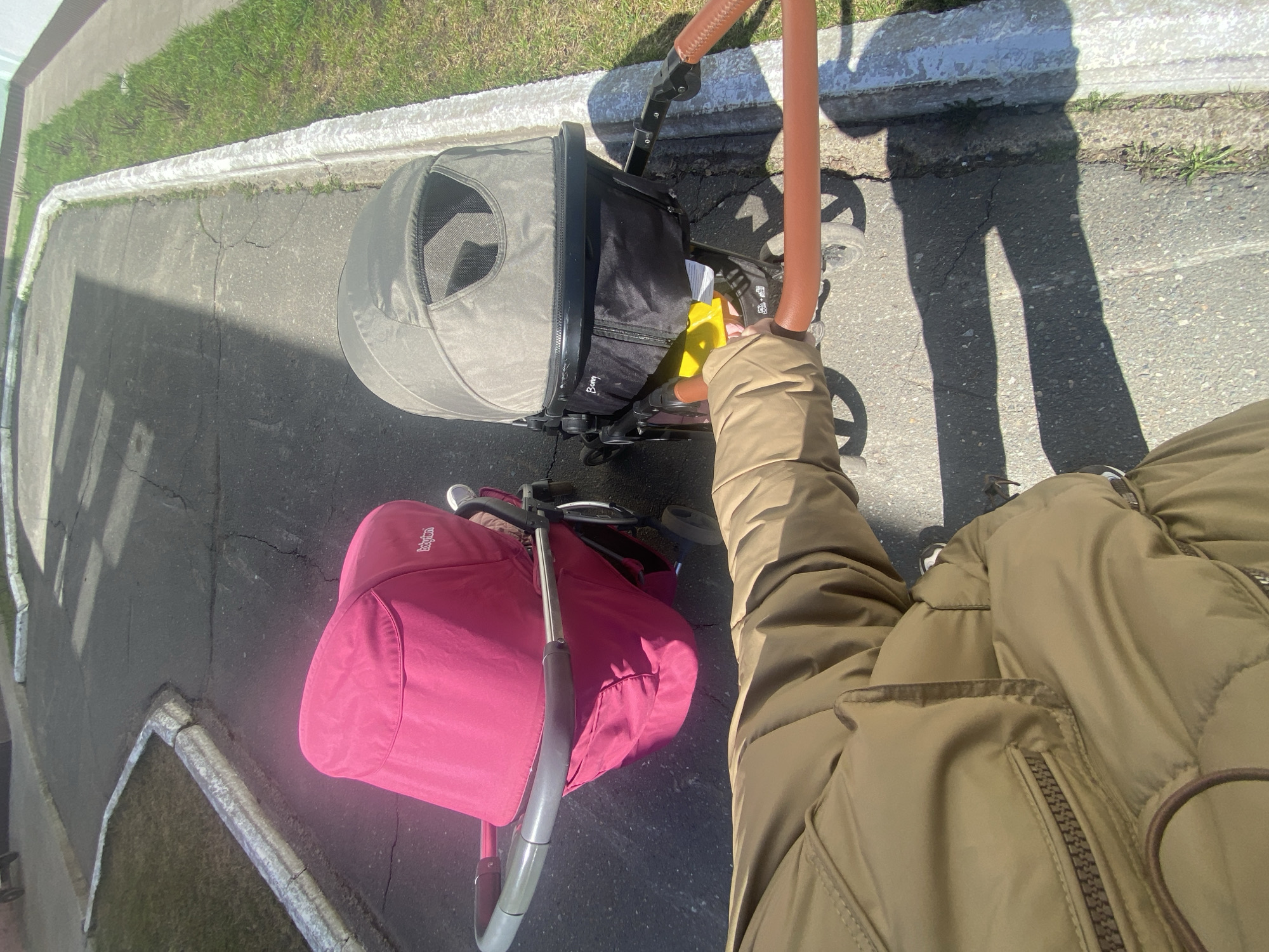 The image shows a person standing on an asphalt surface, likely a sidewalk, next to a curb with grass visible in the background. The scene includes two strollers: one with a gray canopy and black storage underneath, and another with a pink canopy. The person is wearing a brown puffer jacket. The image is taken from a high angle perspective, presumably by the person wearing the jacket, looking down at the strollers. The environment appears to be urban, and based on the prompt, it is located in the city of Chaikovsky, Russia. There are no other people or objects immediately discernible in the image. The interaction is the person is pushing the strollers.