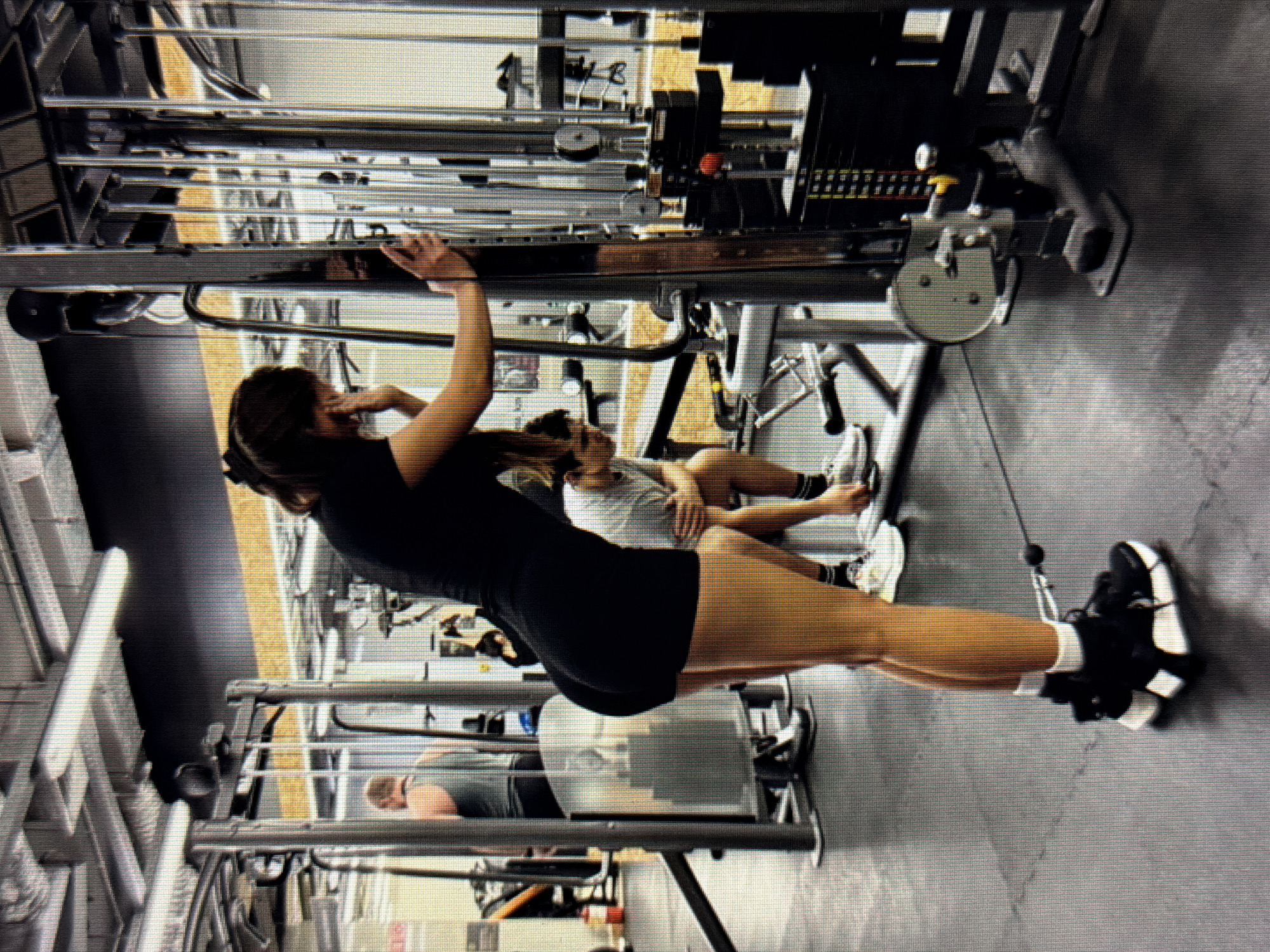 The image depicts an indoor gym environment. In the foreground, a woman wearing a black t-shirt and black shorts is positioned sideways, facing away from the camera and towards a weight machine. She is holding onto the machine with her right hand and her left hand is near her face. A cable is attached to her left ankle. Her hair is pulled back. In the background, another person wearing a grey tank top is visible from the back, standing next to a weight machine. Further back, a person in a grey t-shirt and shorts is seated, looking down. The gym features various weight machines, metal poles, and lighting fixtures on the ceiling. The flooring is dark-colored. The overall scene is consistent with a typical gym setting. The location within the city of Sirius, Russia, is not discernible from the image itself.