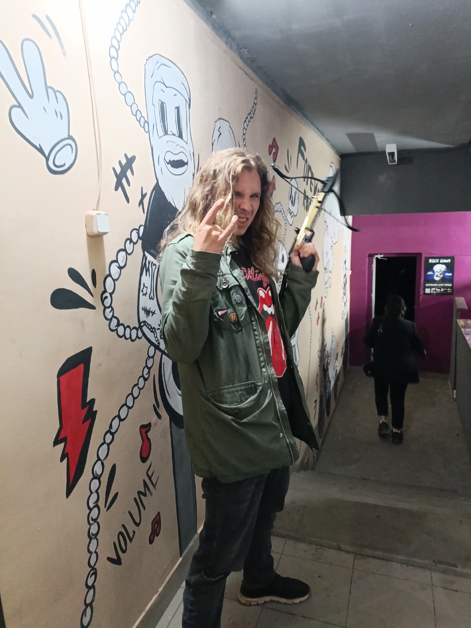 A person with long blond hair and wearing an olive green jacket, a black Rolling Stones t-shirt, and dark jeans stands in a corridor with hand-drawn cartoonish murals on the wall, holding a small crossbow in their right hand. The murals include a hand making a peace sign, a cartoon character, and a lightning bolt with the word "VOLUME". The person is making the "devil horns" hand gesture with their left hand. Behind them, a person in a black jacket walks towards a doorway leading into a magenta-colored room, where a poster or sign is visible on the wall. The scene appears to be inside a building, possibly a music venue or a recreation space. The location is identified as the city of Chaikovsky, Russia.