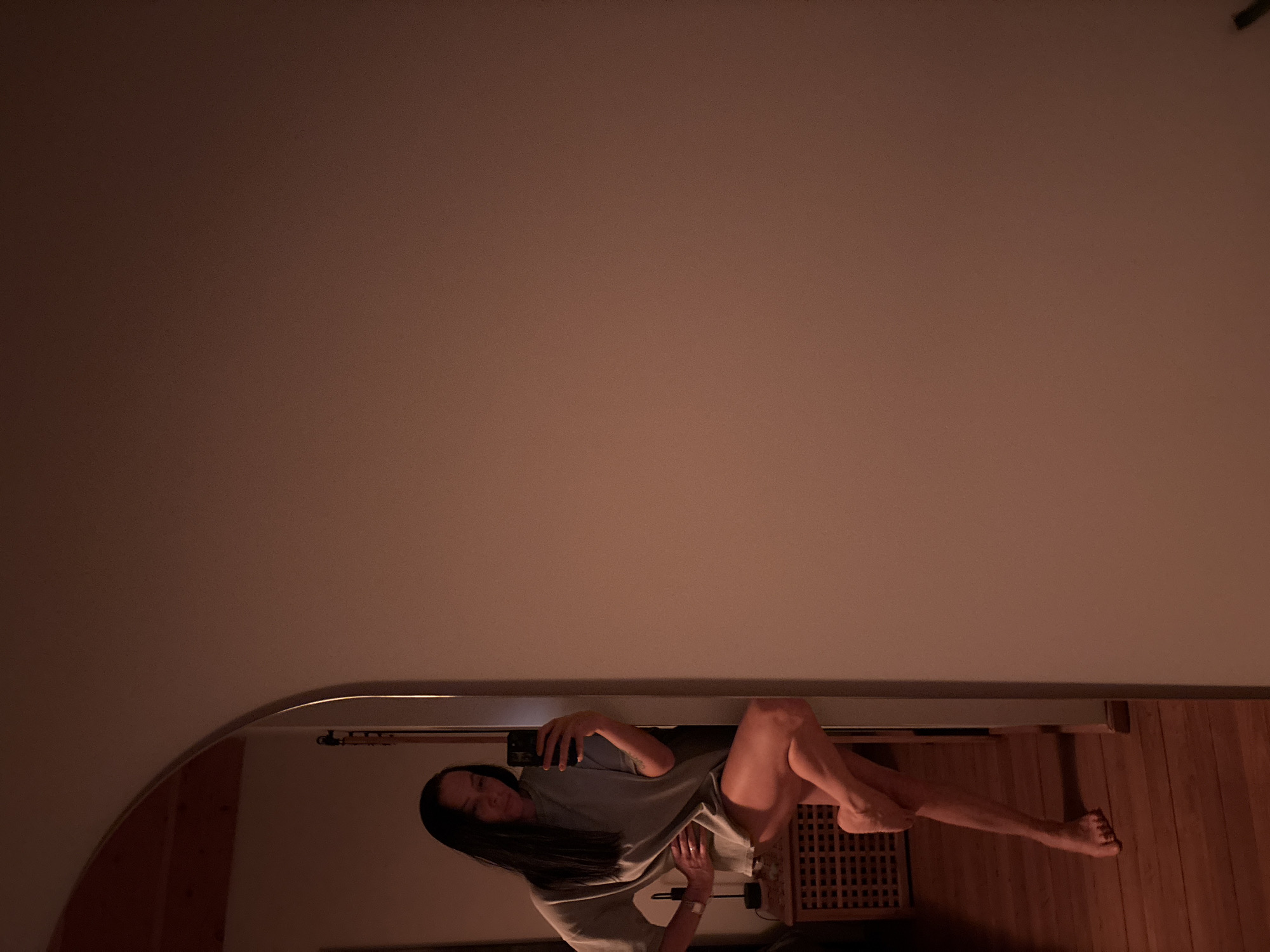 The image displays an indoor residential scene, likely within the city of Chaikovsky, Russia; however, the precise location within the city is not identifiable from the visual evidence. A female individual with long dark hair is visible in a mirror reflection, holding a black smartphone to take a selfie. She is wearing an oversized, light-colored t-shirt that extends to mid-thigh, and she is barefoot. Her left hand rests on her hip, while her right leg is bent at the knee and crossed in front of her left leg, with her right foot touching the floor. The mirror features an arched top and is positioned within what appears to be a doorway or architectural arch. To the left, reflected in the mirror, is a dark wooden cabinet or table with a slatted front, upon which a black table lamp with a dark shade is placed. The flooring consists of wooden planks. The wall to the immediate right of the mirror is a plain, light brown or peach color. The environment is dimly lit with warm, artificial light.