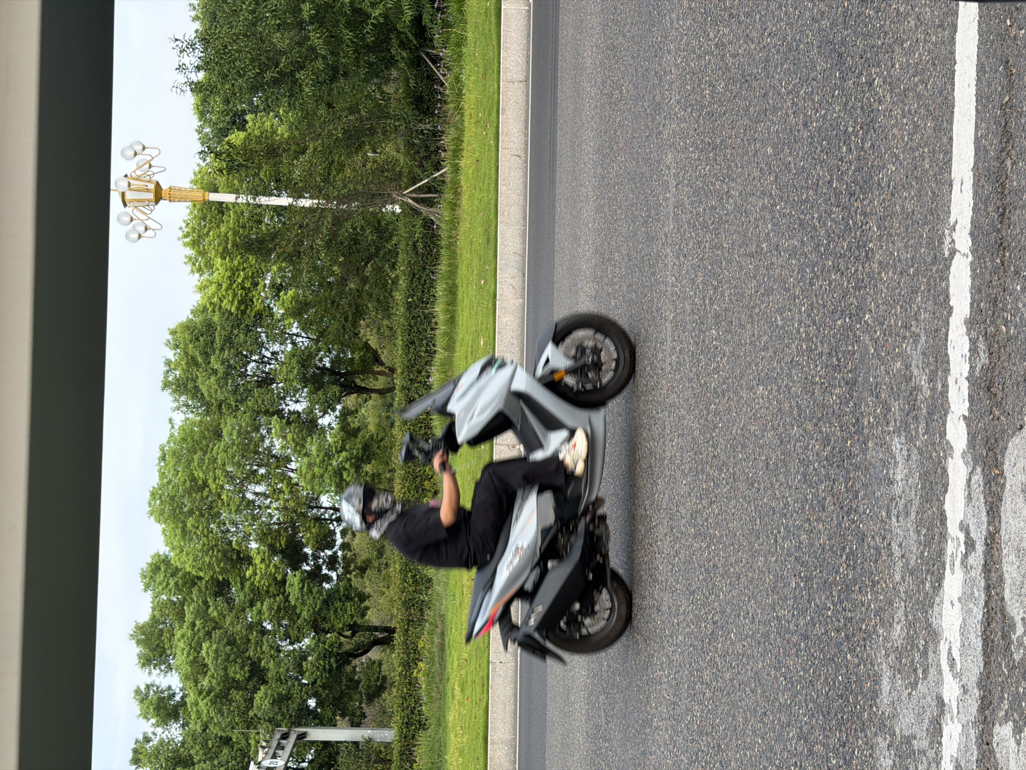 A person wearing a black shirt, black pants, and a patterned helmet is riding a light gray scooter on a paved road. The scooter has a black exhaust pipe and a dark seat. The person's right foot is visible, wearing a beige sneaker. The background consists of a grassy median with trees behind it. A decorative lamppost with multiple light bulbs is visible to the right of the scooter. The image appears to have been taken from a slightly elevated position, possibly from a vehicle or elevated walkway. There is a white line painted on the road in the foreground. The location is identified as Jiangjin, China.