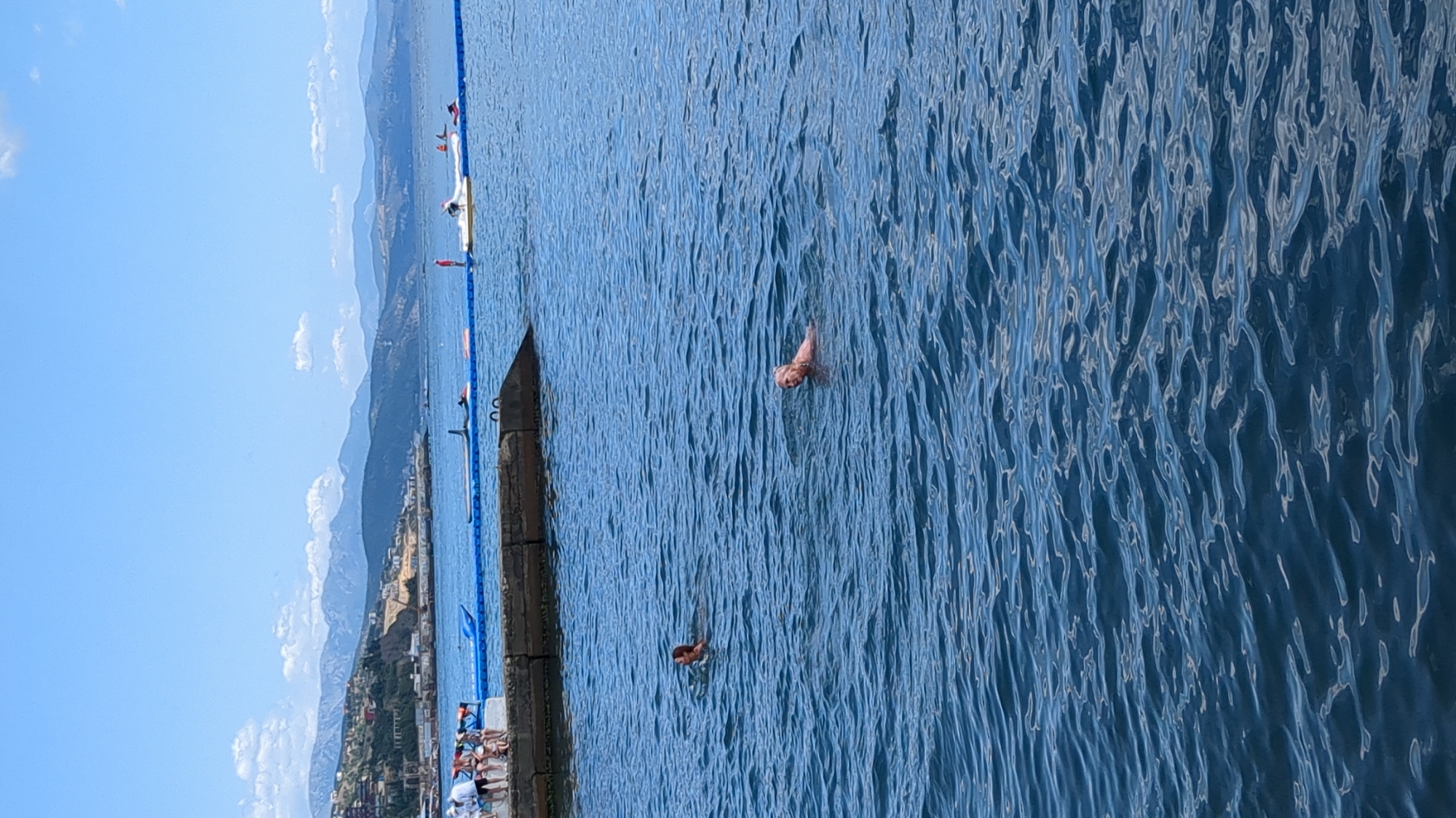 The image depicts a body of water, likely a sea, on a clear, sunny day with a hazy blue sky. In the foreground, the water is a deep blue with gentle ripples. Two individuals are swimming in the water, both appearing to be adults. The person on the right is closer to the viewer and is looking towards the camera, with their upper body visible. They have light-colored hair. The person on the left is further away and is swimming away from the camera, with only their head and shoulders visible.
In the mid-ground, a concrete pier or seawall extends into the water from the left side of the image. Several people are gathered on this structure, appearing to be dressed in casual clothing, some wearing life vests. Beyond the pier, a blue inflatable barrier or boom is visible in the water, delineating a swimming area. Several small watercraft, including what appear to be jet skis and a stand-up paddleboard, are present near the barrier.
In the background, a coastline is visible, characterized by hills or mountains with sparse vegetation and some buildings. The landscape suggests a coastal town or city. The distant mountains are shrouded in a light haze, consistent with a warm climate. The overall scene is one of recreational activity on a body of water, with a backdrop of a coastal landscape. The location is identified as Alushta, Ukraine.