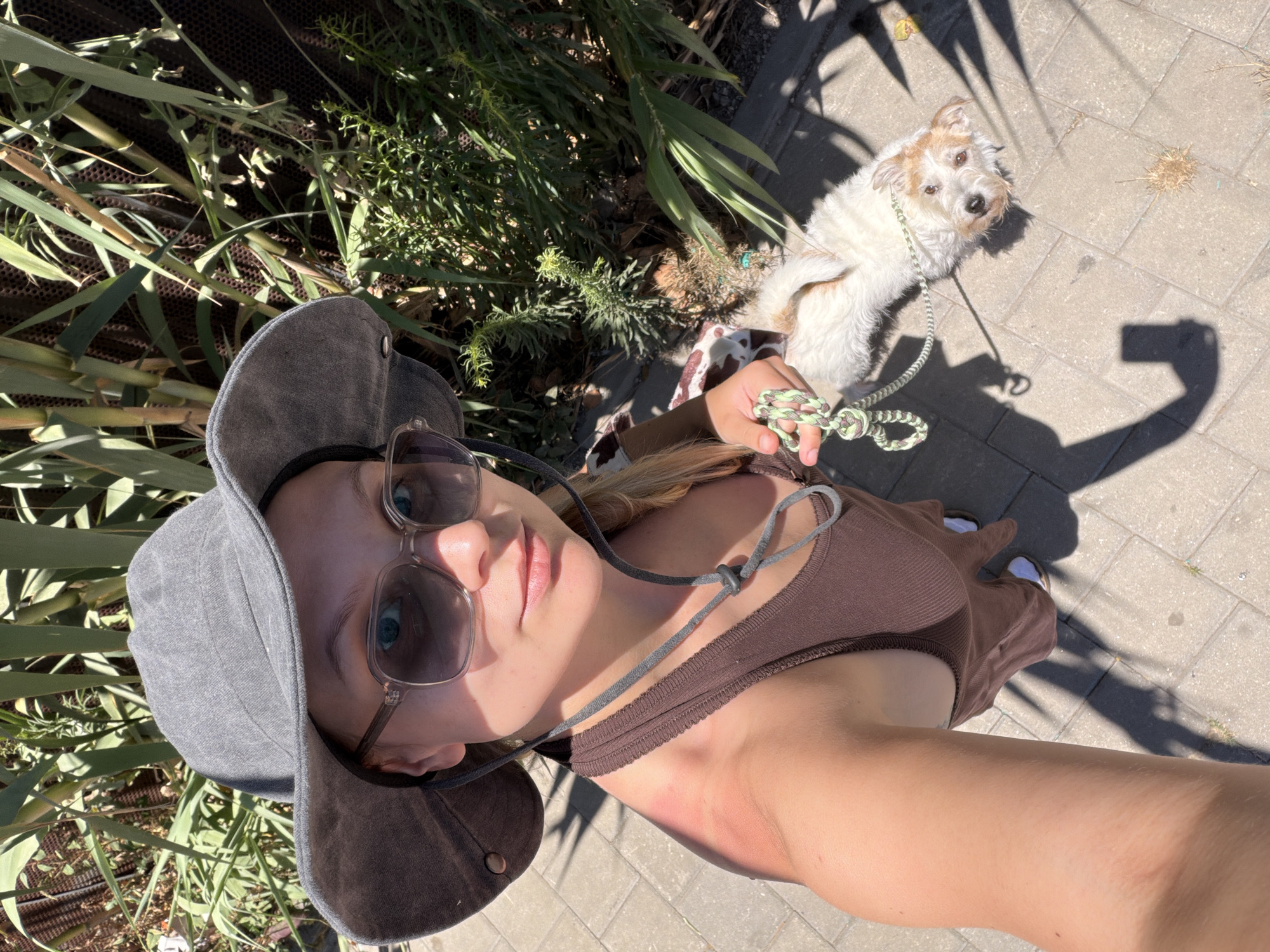A first-person perspective selfie captures a person and their dog outdoors on a sunny day in Tel-Aviv, Israel. The person, appearing to be female, is in the foreground, angled upwards from the chest. She is wearing a grey bucket hat and mirrored sunglasses that reveal her bright blue eyes. She is dressed in a brown ribbed tank top or dress, and her right arm, showing a faint reddish mark on the shoulder, extends into the frame, holding a distinctive green and white braided dog leash.
Beside her, on a light-colored paved sidewalk made of rectangular blocks, stands a small, scruffy white dog with light brown patches, possibly a terrier mix. The dog is looking directly up towards the camera with an alert and inquisitive expression. Its leash is clearly visible, connecting it to the person's hand.
The scene is bathed in bright sunlight, casting sharp shadows on the pavement, including the recognizable shadow of a phone or camera, indicating a selfie. Dense green foliage and plants line the left side of the frame, suggesting a park or a green urban area. The activity appears to be a casual walk or outdoor outing. The time of day is clearly bright, likely midday or early afternoon, with clear weather. No visible text is present in the image.